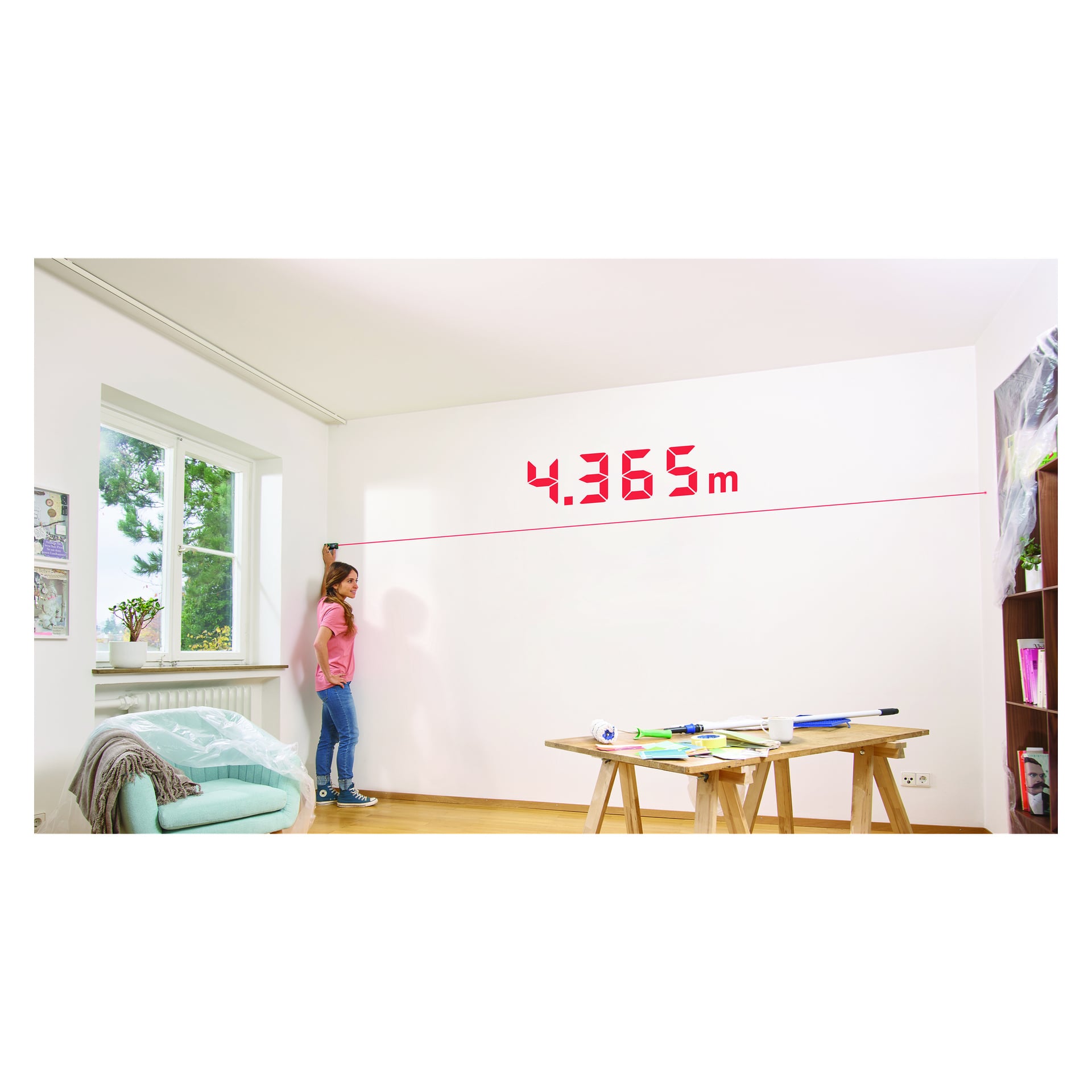 Osoba mierzy odległość za pomocą miernika odległości Bosch EasyDistance 20, który na ścianie pokazuje 4,365 m na czerwono. W pomieszczeniu znajduje się okno, półka, stół przykryty narzędziami oraz krzesło owinięte w plastik.