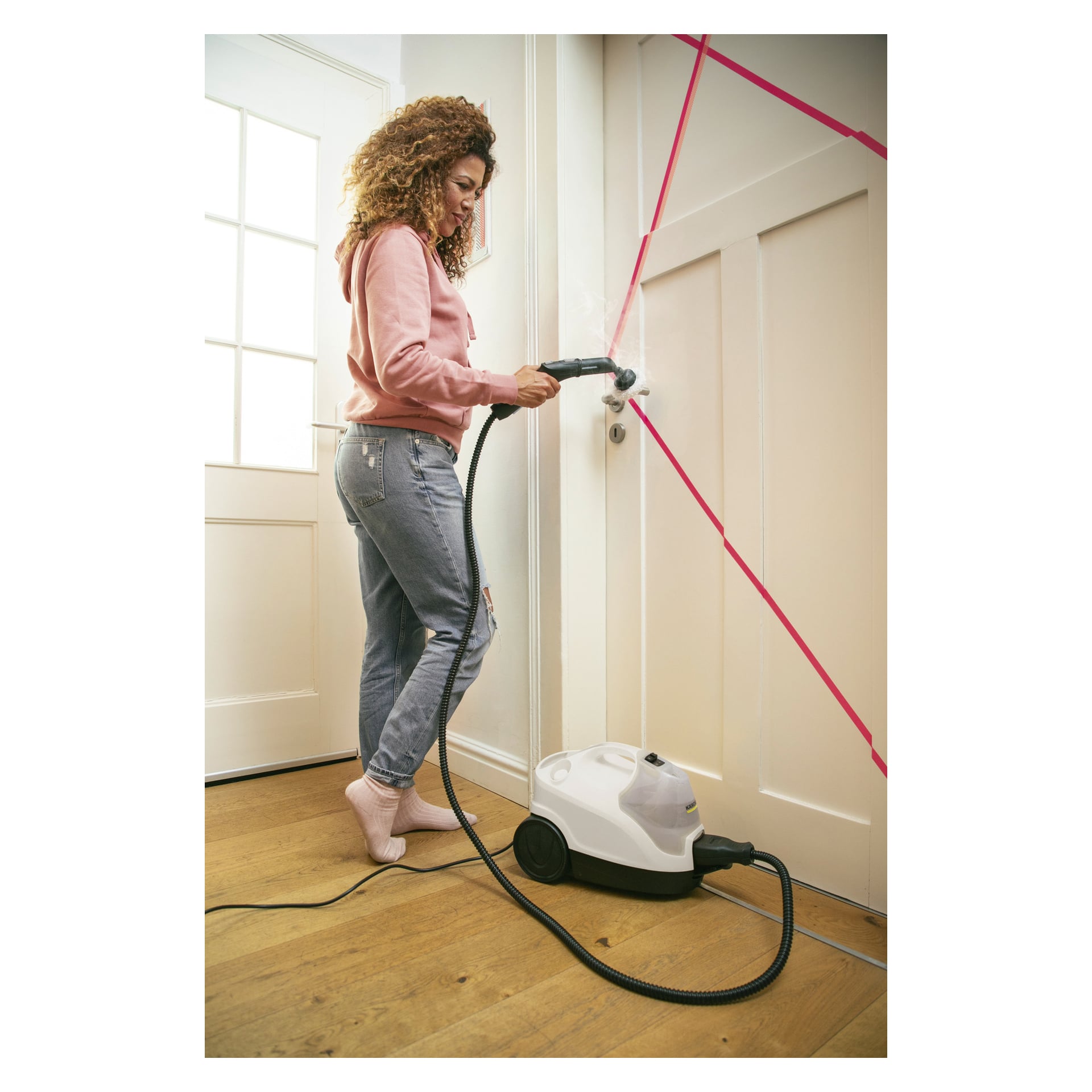 En kvinna i en rosa hoodie rengör en vit dörr märkt med ett rosa tejp "X" med hjälp av Kärcher Steam Cleaner SC 4 EasyFix 9-delat set i ett väl upplyst rum med trägolv.