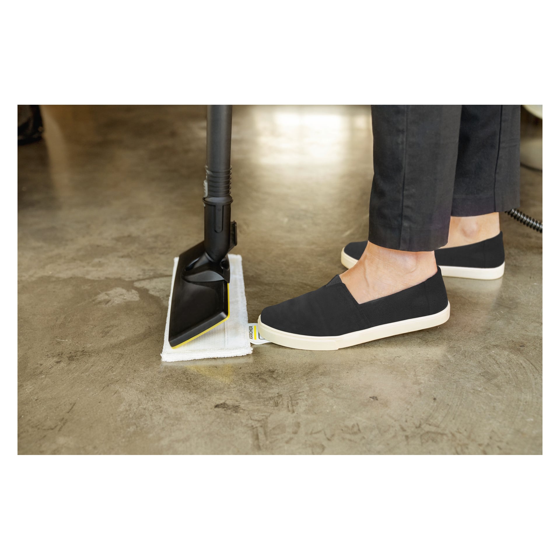 Någon i mörka byxor och svarta slip-on-skor använder foten för att fästa en rengöringsdyna på Kärcher Steam Cleaner SC 4 EasyFix 9-delat set, redo att rengöra ett slätt betonggolv.