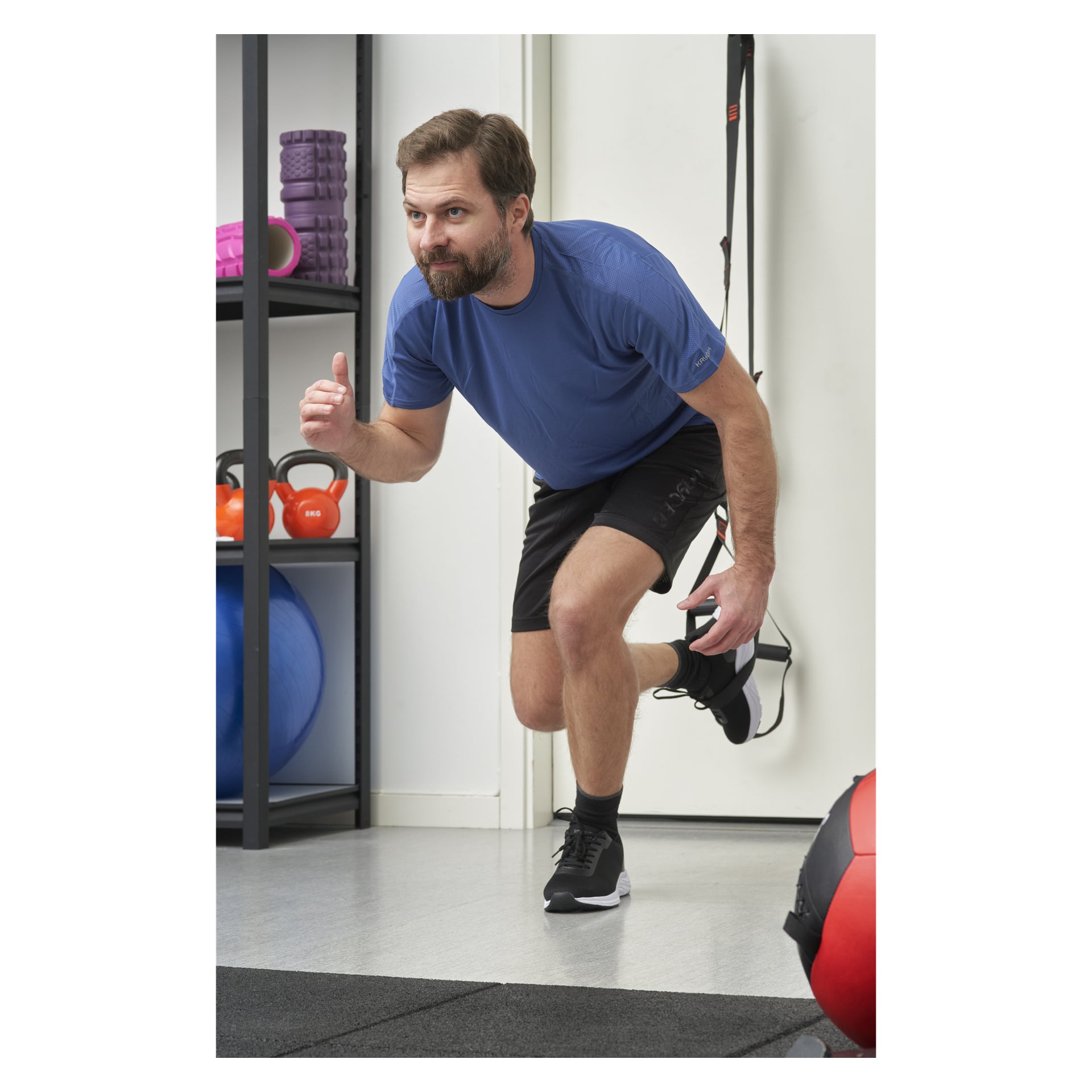 En skäggig man med kort hår står på ett ben i ett gym, klädd i blå skjorta, svarta shorts och sneakers. Hyllor bakom honom innehåller Kayoba-träningsband, kettlebells, en stabilitetsboll och annan gymutrustning.