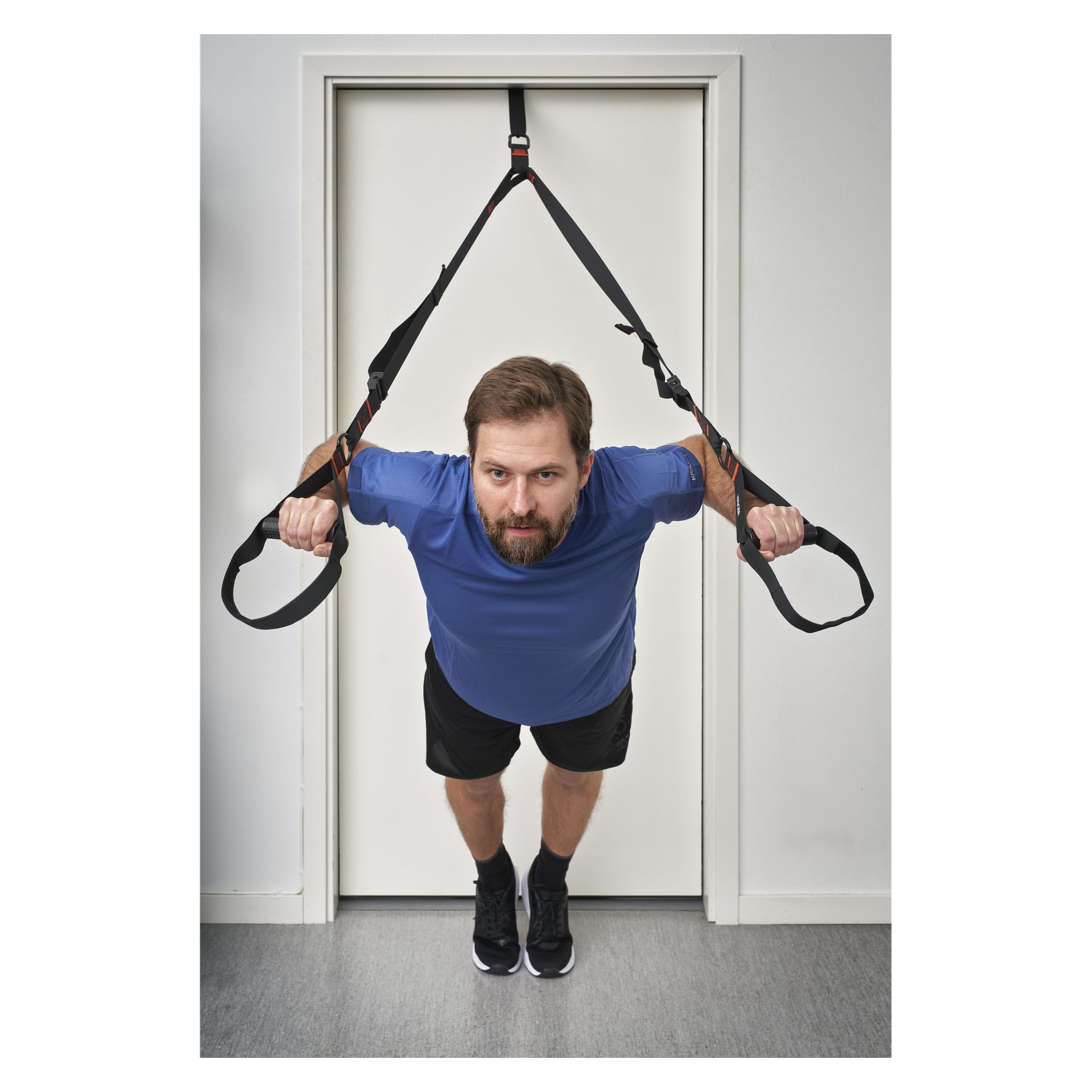 En skäggig man tränar inomhus med Kayoba Exercise Bands fästa vid en dörr och utför en lutande armhävning. Han är klädd i en blå t-shirt, svarta shorts och svarta sneakers, med en vit dörr synlig i bakgrunden.