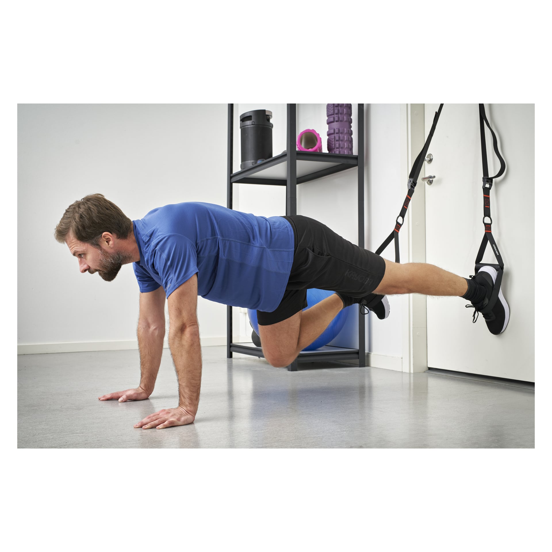 En man gör en upphängd planka, hans fötter fastsatta i Kayoba Exercise-band fästa vid en dörr. Han har på sig en blå skjorta och svarta shorts, medan extra träningskläder syns på en hylla bakom honom.