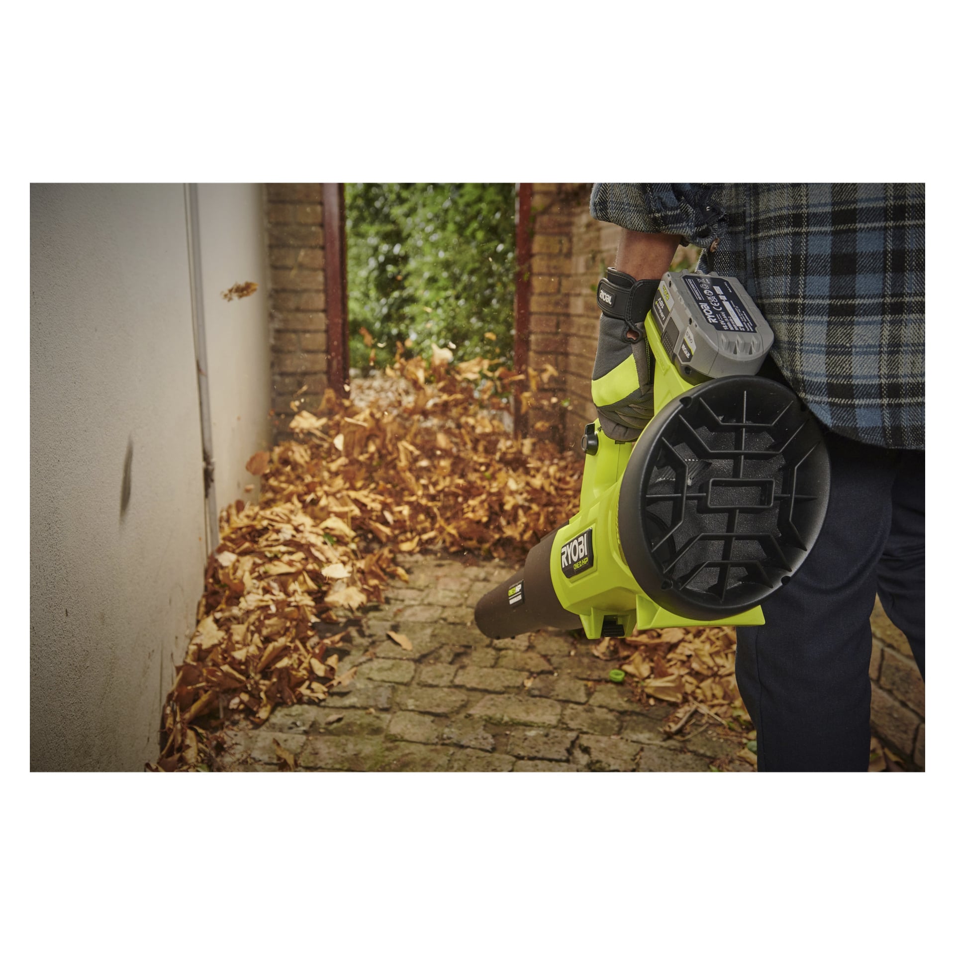 En person med handskar och rutig skjorta kör den batteridrivna lövblåsaren RY18BLXD-0 i Ryobi One+ HP, där torra löv rensas bort från en stenstig omgiven av en vit vägg på ena sidan och en tegelvägg på den andra, med växter synliga längre fram längs gången.
