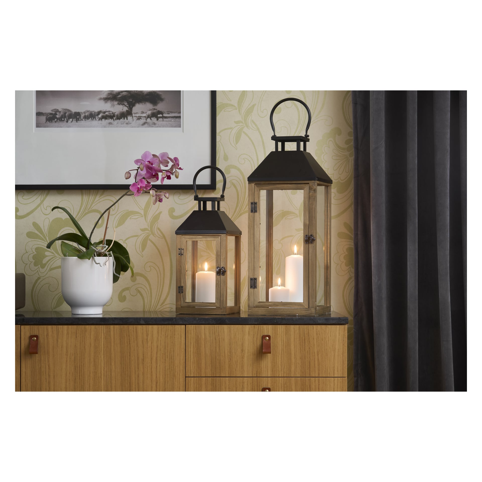 Ett träskåp visar en vit orkidé i en kruka och två EKVIP-lyktor med lysande ljus inuti. Ovanför hänger ett inramat svartvitt foto, med mörka gardiner till höger.