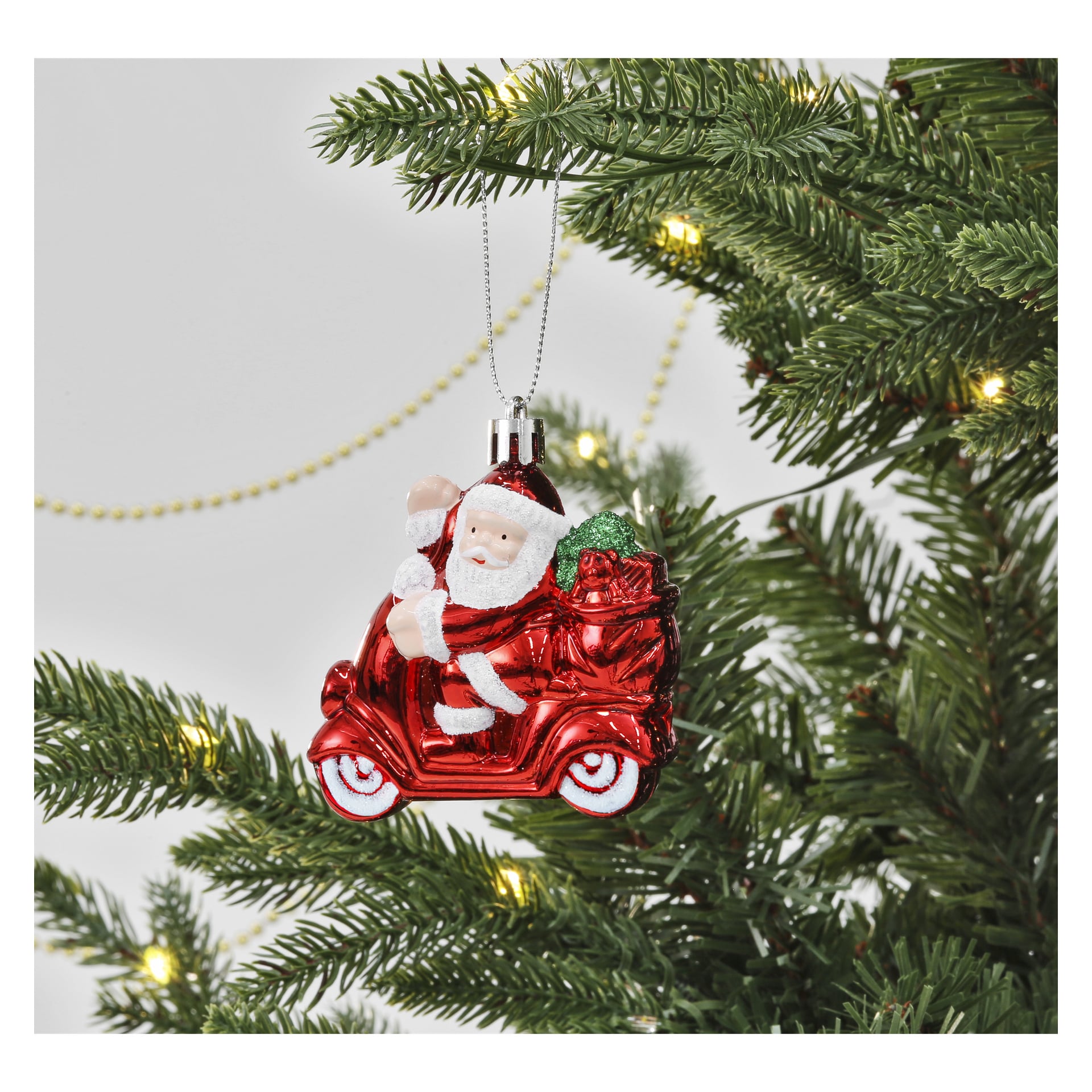 En jultomte-prydnad från EKVIP:s tredelade julgransdekorationsset, som visas åka en röd scooter med en påse fulla av presenter, hänger på en grön julgranskvist prydd med vita ljus och guldpärlgirlander i bakgrunden.