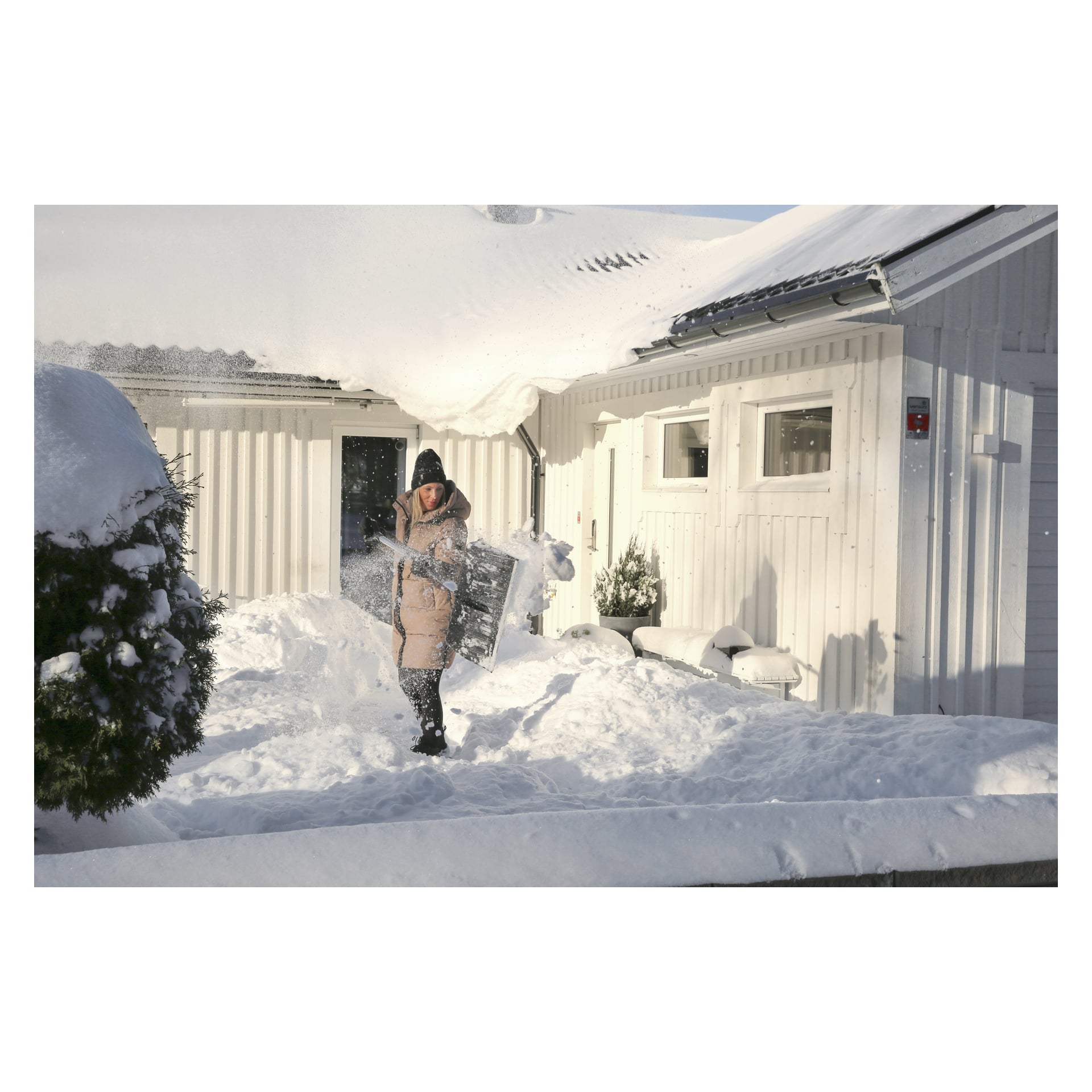 En person klädd i vinterkläder använder en Hard Head snöskyffel med aluminiumhandtag för att ta bort snö utanför ett vitt hus. Snö täcker marken, taket och buskarna runt omgivande. Scenen är upplyst av solljus under en klar himmel.