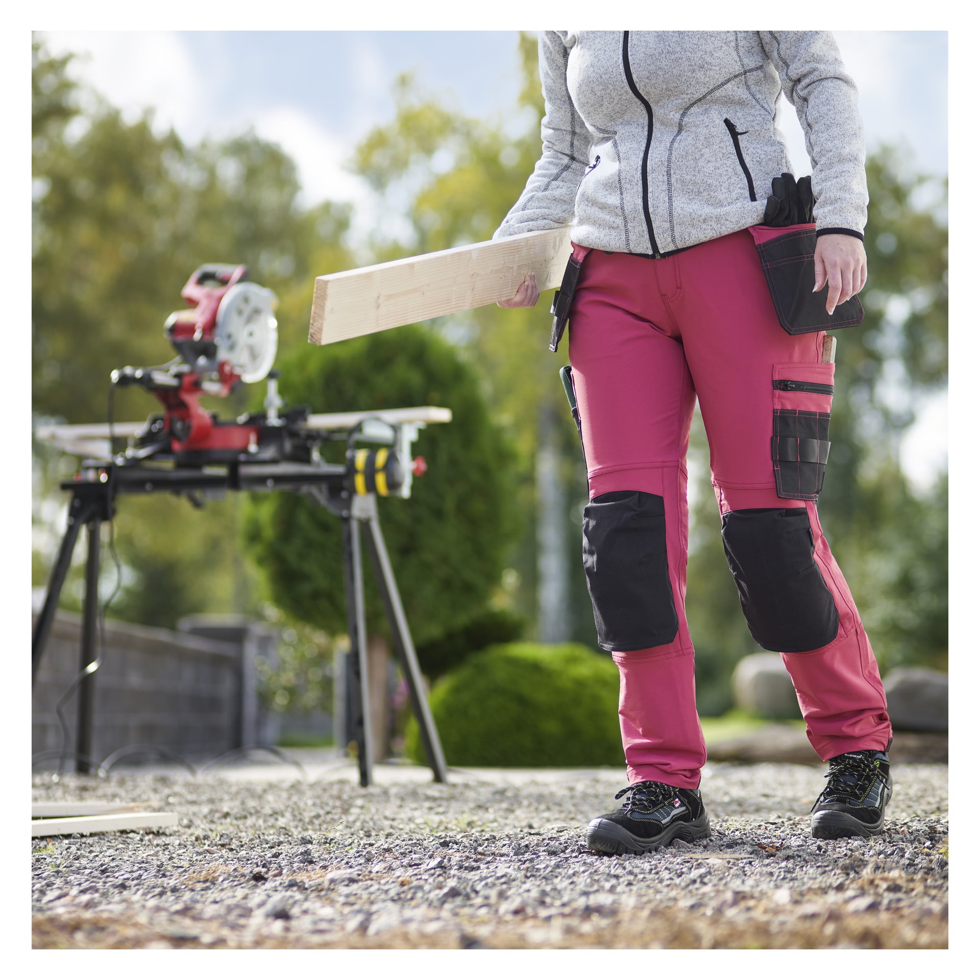 En person som bär röda Bluewear Pro Workwear Women's arbetsbyxor med knäskydd står utanför vid en geringssåg och håller i en träplanka. Träd och buskar syns i bakgrunden.