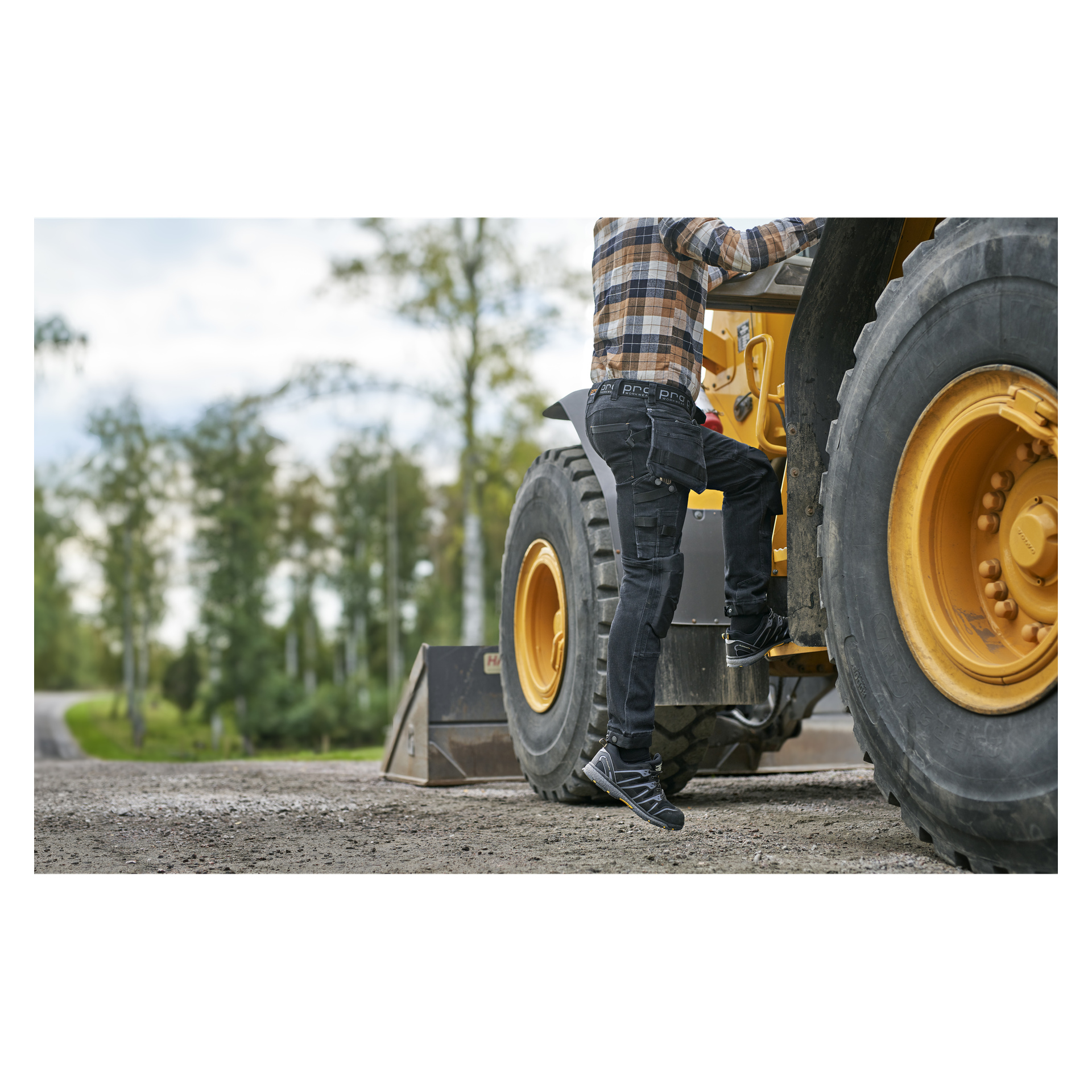 Någon klädd i en rutig skjorta, Bluewear Pro Workwear herrbyxor från Steve Denim och sneakers kliver in i ett stort gult byggfordon parkerat på en grusväg, omgiven av träd och frodig grönska.