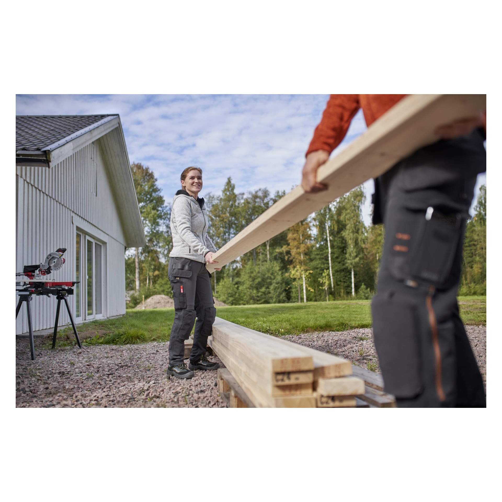 Två personer i arbetskläder bär en lång planka utanför ett hus. En ler mot kameran och har på sig Bluewear Pro Workwear Women's arbetsbyxor Stella. Staplar av trä och en elektrisk såg på ett stativ finns i närheten.
