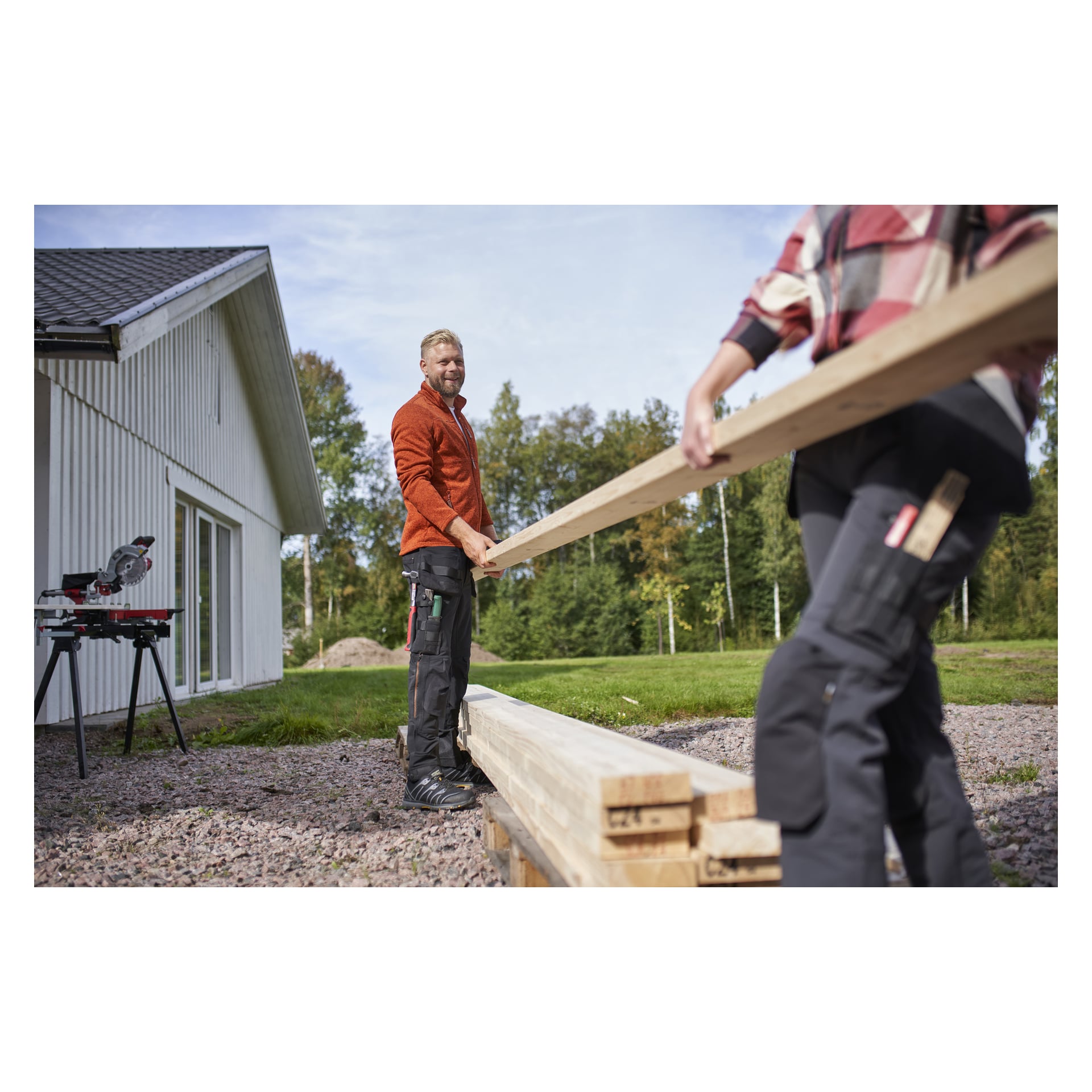 Två personer bär träbrädor. En, klädd i herrbyxor Steve från Bluewear Pro Workwear, vänder sig mot kameran och ler; en såg och andra verktyg finns i bakgrunden.