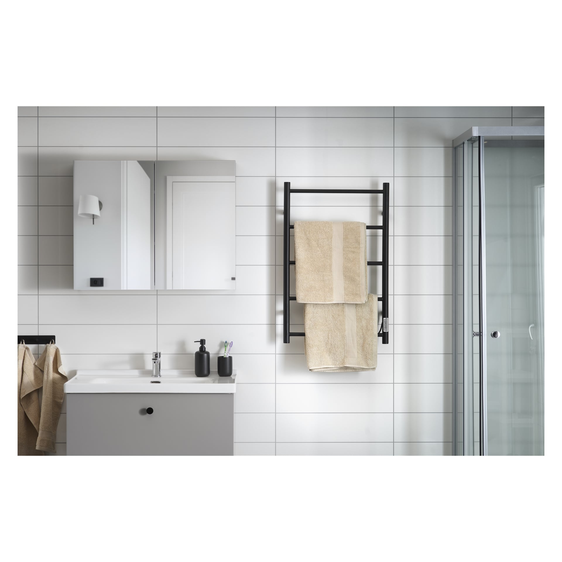 Modernt badrum med vitt kakel, dusch i glas, spegelskåp, handdukshängare med beige handdukar, grått handfat och svarta dispensrar.
