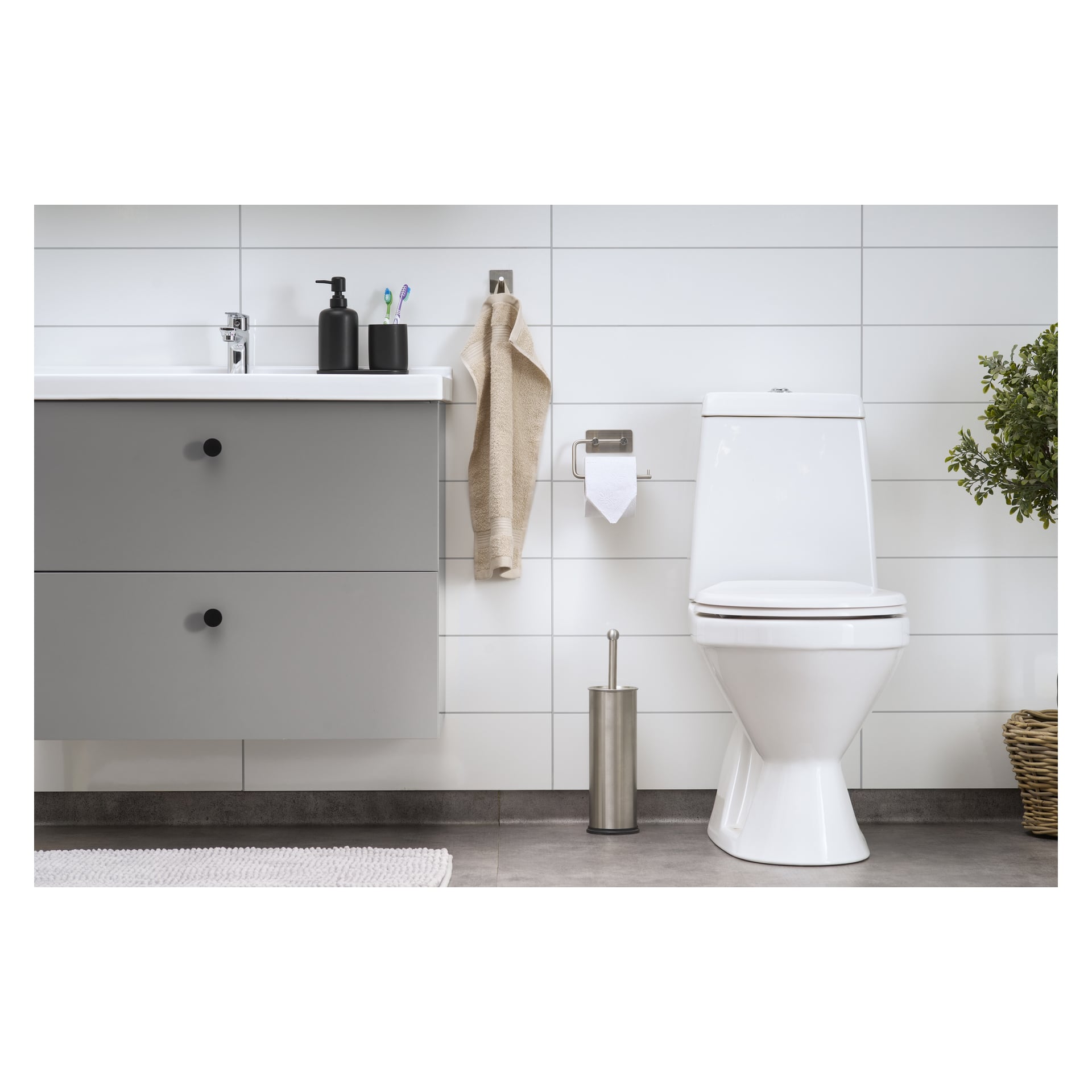 Ett modernt badrum med en vit toalett, ett grått sminkbord, en handduk hängande på en krok, toalettpapper på sin hållare, en svart tvålbehållare, tandborstar samlade i en kopp, EKVIP-toalettborsten i närheten, en krukväxt som tillför grönska och en tunn matta som täcker det grå kaklade golvet.