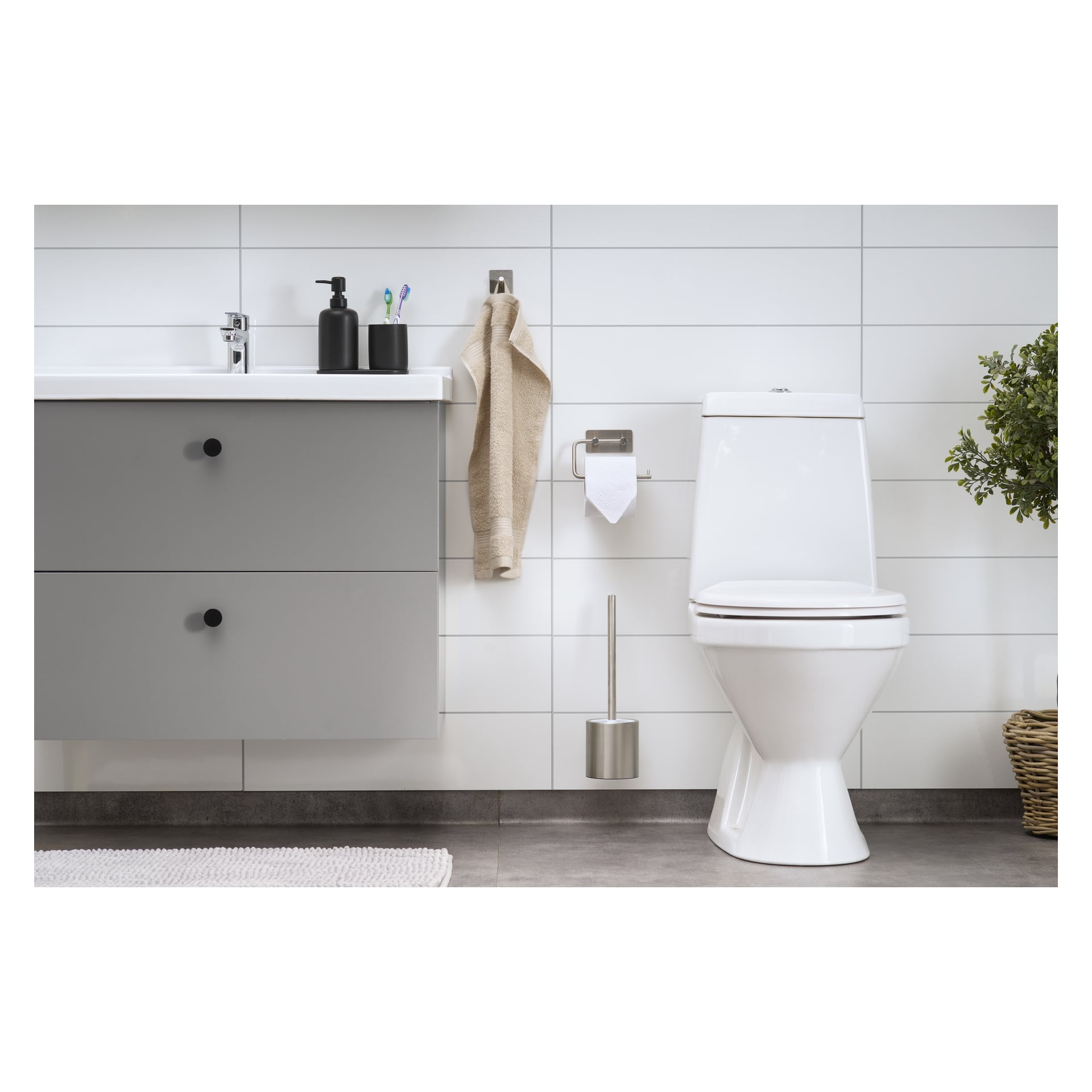Modern badrum med EKVIP toalettborste, vit toalett, monterad toalettpappershållare, grå kommoditet accentuerad av svarta handtag, handfatsområde utrustat med tvåldispenser och tandborstar, beige handduk, grön krukväxt och vit kaklade vägg.