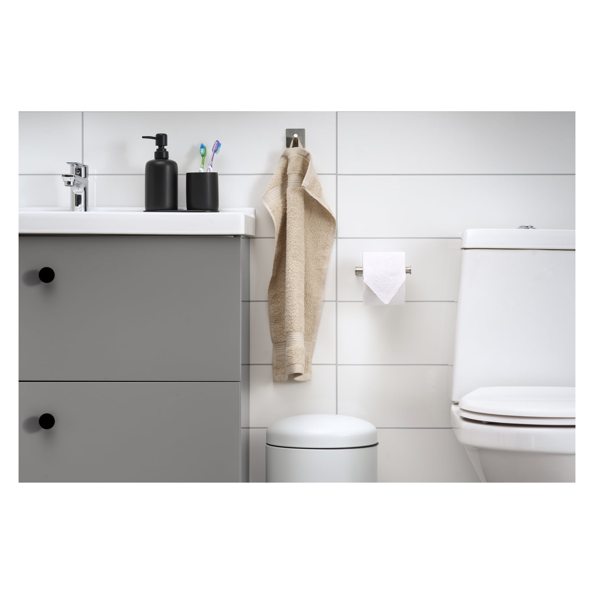 Ett stilrent badrum med ett grått skåp, två tandborstar i en svart hållare, en tvålautomat, en beige handduk som hänger upp, en vit toalett, en EKVIP toalettpappershållare och en liten soptunna längs vita kaklade väggar.