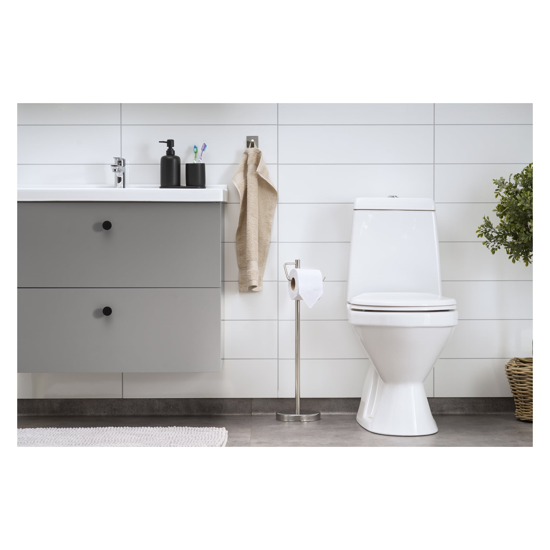 Ett modernt badrum med vit toalett, grå sminkbord med handfat och olika toalettartiklar, en beige handduk på en väggkrok, EKVIP toalettpappershållare installerad i närheten, en grön krukväxt som dekoration och väggar täckta med vita kakelplattor.