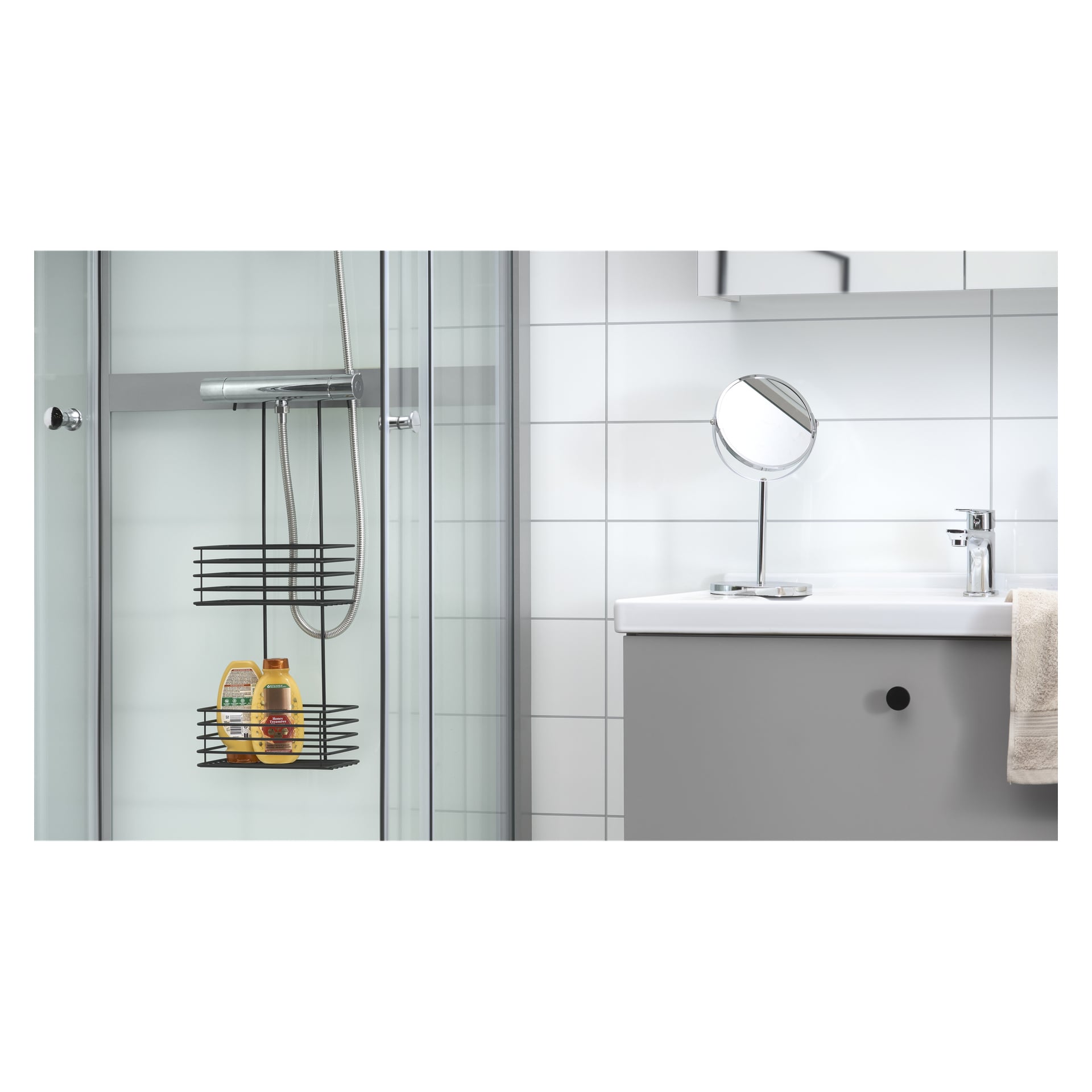 Ein Badezimmer mit Dusche mit Glastür, einem EKVIP Black-Duschregal mit zwei Flaschen, einem modernen Waschbecken mit einem darüber hängenden Handtuch, einem runden Spiegel über dem Waschbecken und Wänden mit weißen Fliesen.