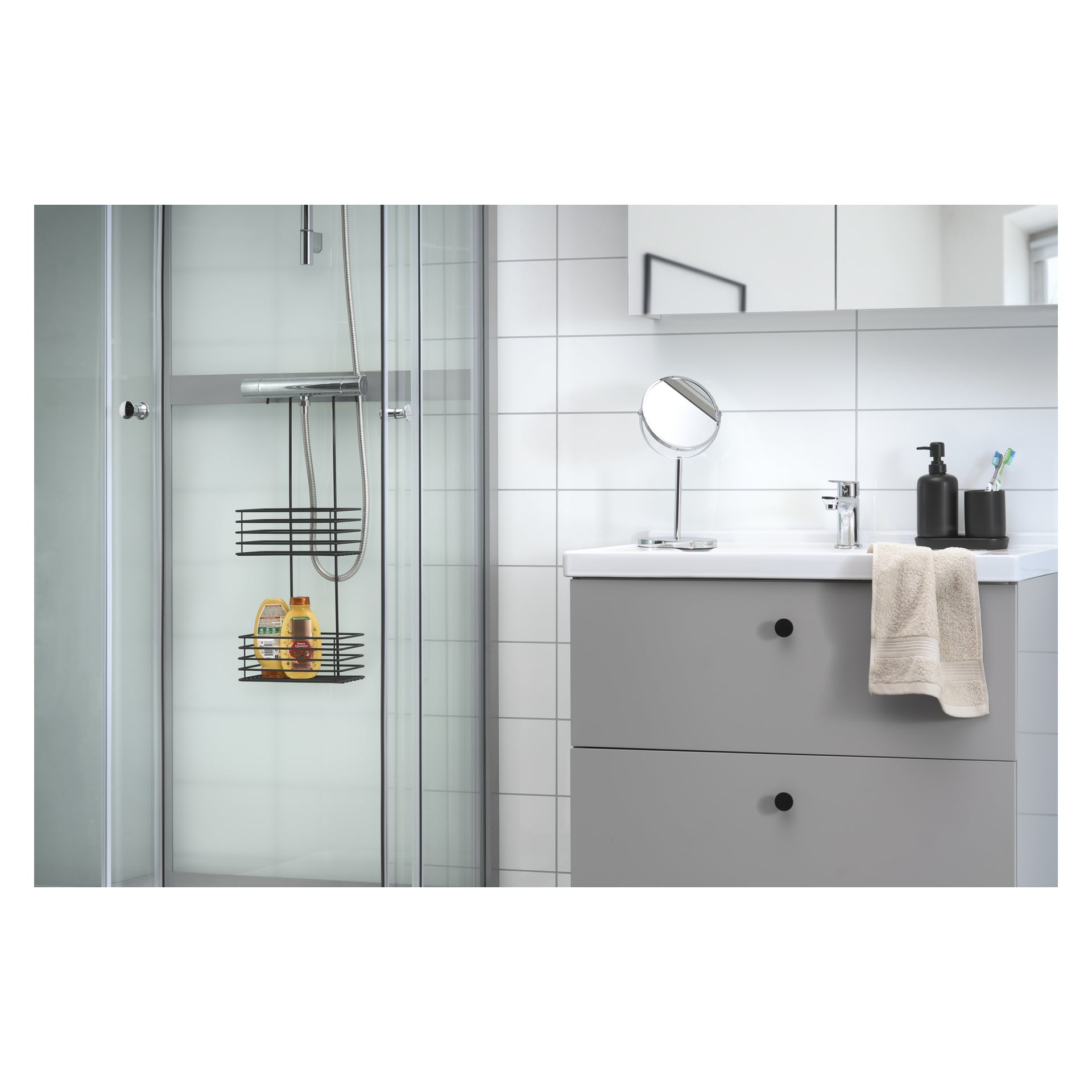 Ein elegantes Badezimmer mit einer Glasduschkabine, weiß gefliesten Wänden und einem grauen Waschtisch mit einem Handtuch, einem Seifenspender und einem Spiegel obendrauf. Im Inneren der Dusche sind zwei Flaschen mit Duschprodukten auf einer EKVIP Black Shower Shelf Unit organisiert.