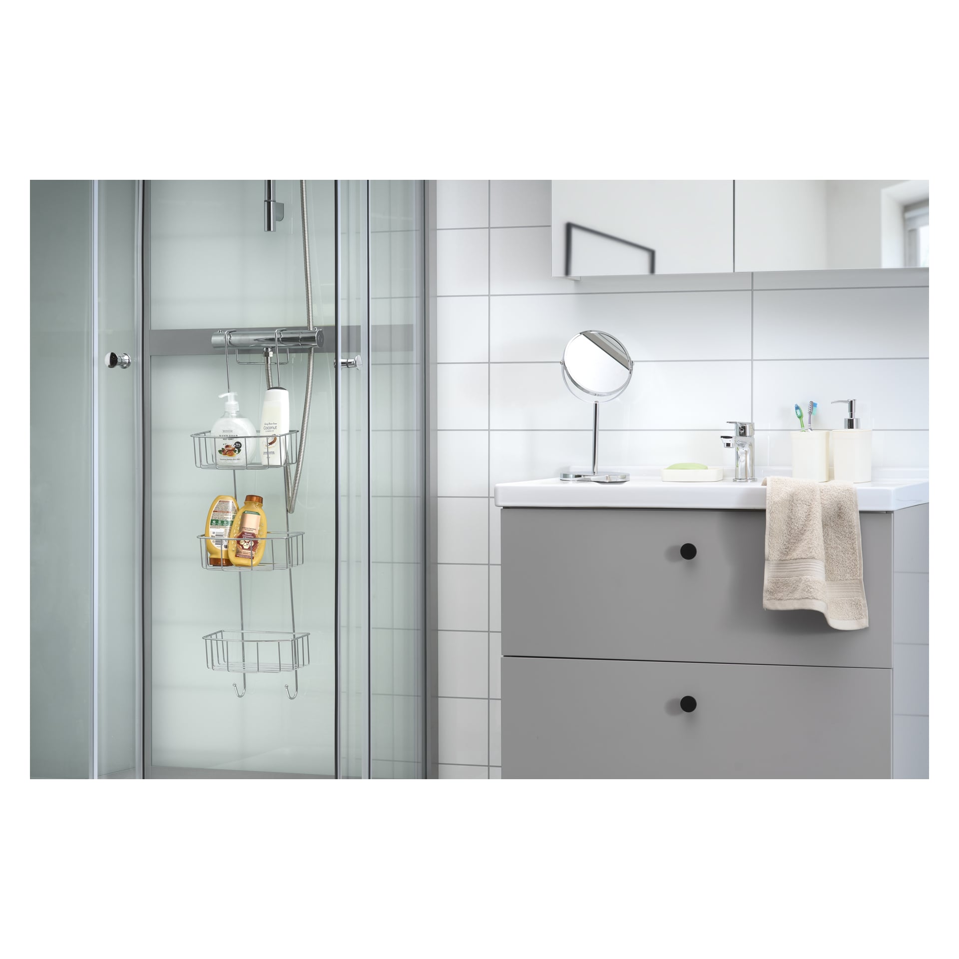 Ett modernt badrum har en glasinglasad dusch, grå sminkbord och en rund spegel, accentuerad av slanka tvåldispensrar, tandborstar och en handduk. EKVIP Shower-hyllan organiserar tvålar och flaskor inne i duschen. Vita kakelväggar och riklig belysning skapar en fräsch atmosfär.