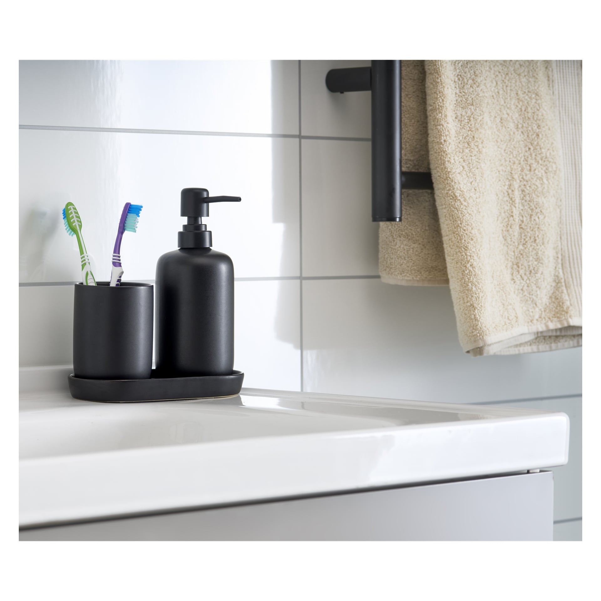 Ett tredelat EKVIP Soap Dispenser Set, med en svart tvåldispenser och en tandborsthållare med två tandborstar, står på en badrumshandfat. I bakgrunden hänger beige handdukar över ett svart handduksställ.