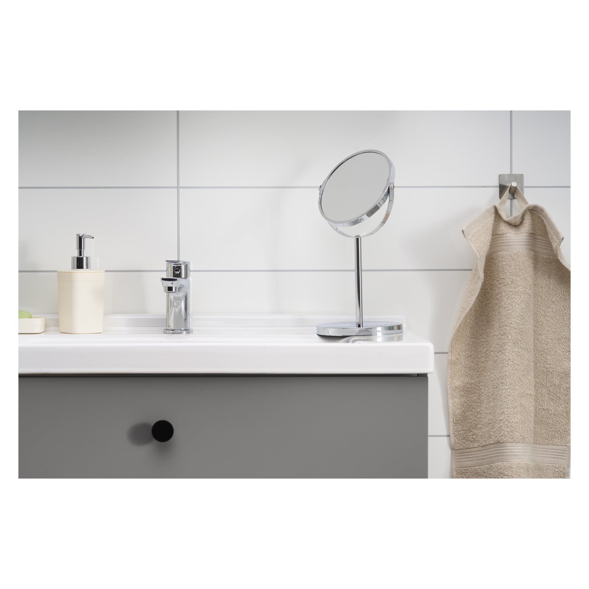 En slank badrumsbänk visar EKVAP-spegeln, en tvåldispenser, en kran och en beige handduk draperad på en väggkrok. Vita kakel utgör bakgrunden, och en tvålbit vilar på vänster sida.