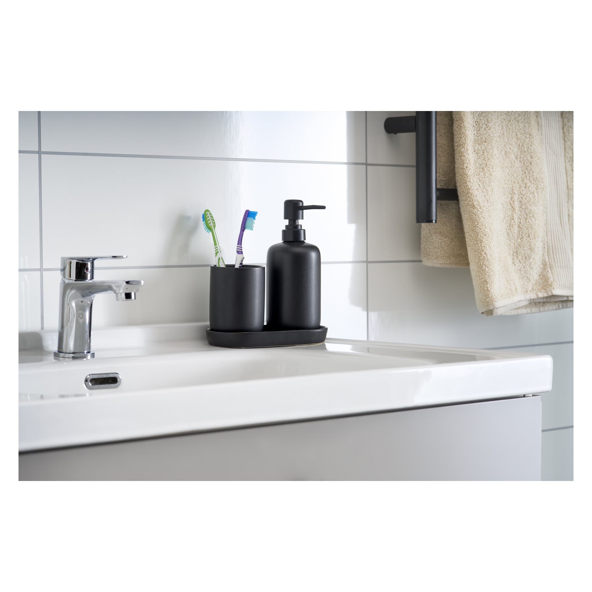 En vit handfat med silverkran rymmer den tredelade EKVIP Soap dispensern i svart, med en tvålbehållare och en tandborsthållare med två tandborstar. En beige handduk hänger över en ställning i närheten.
