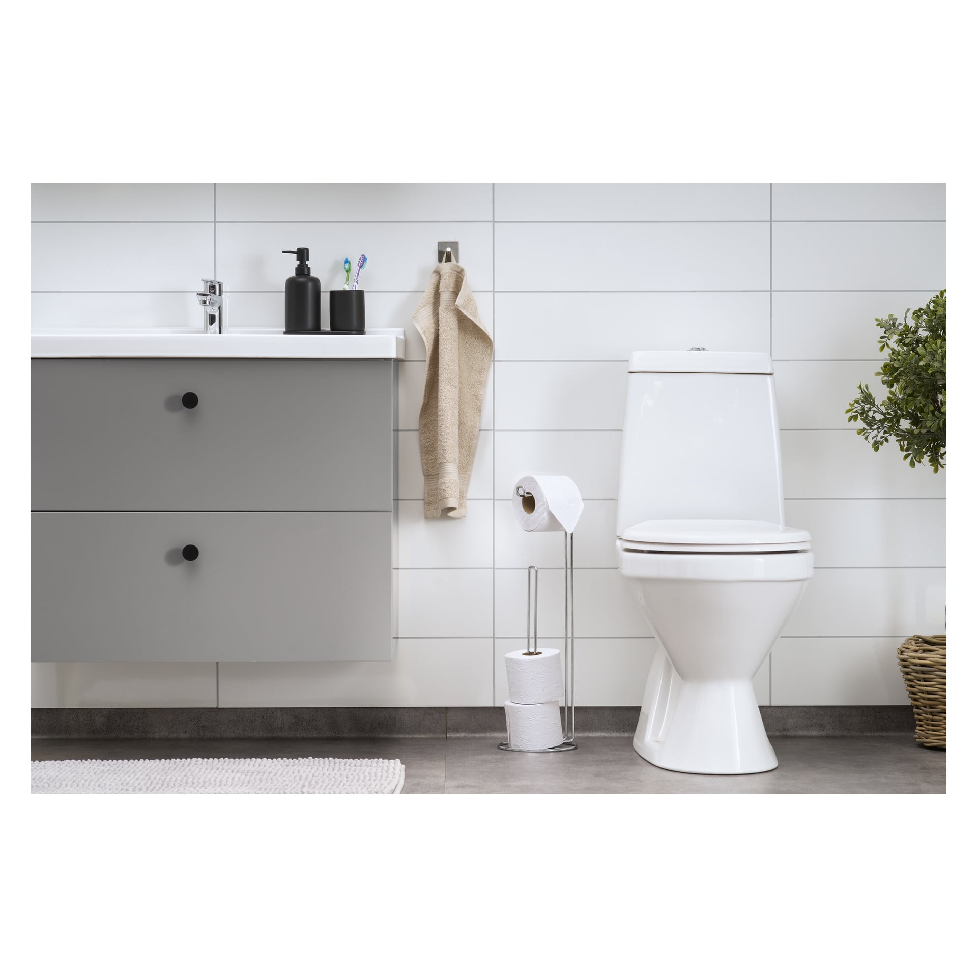 Ett modernt badrum med vit toalett, EKVIP 2-i-1 toalettpappershållare och en grå sminkbord med handfat, tvålautomat och flera tandborstar ovanpå. En beige handduk hänger på väggen, och en grön växt i en vävd korg vilar på golvet.