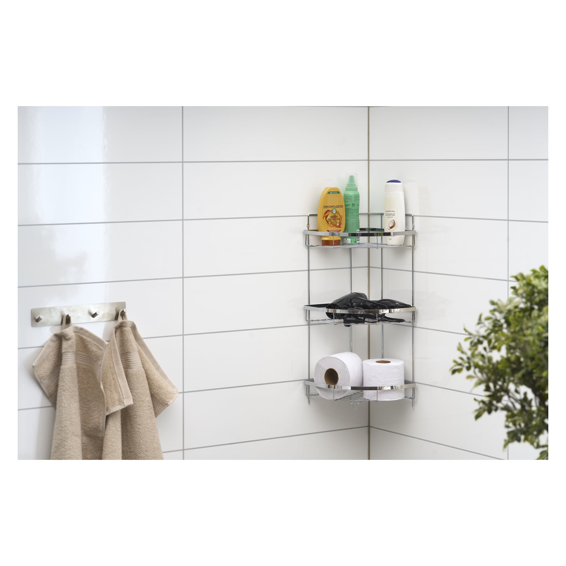 En EKVAP duschhylla är fylld med schampo, balsam, duschtvål, en svart hårtork och två toarullar. I närheten hänger två beige handdukar, och en grön växt dyker upp i nedre högra hörnet mot kaklade väggar.