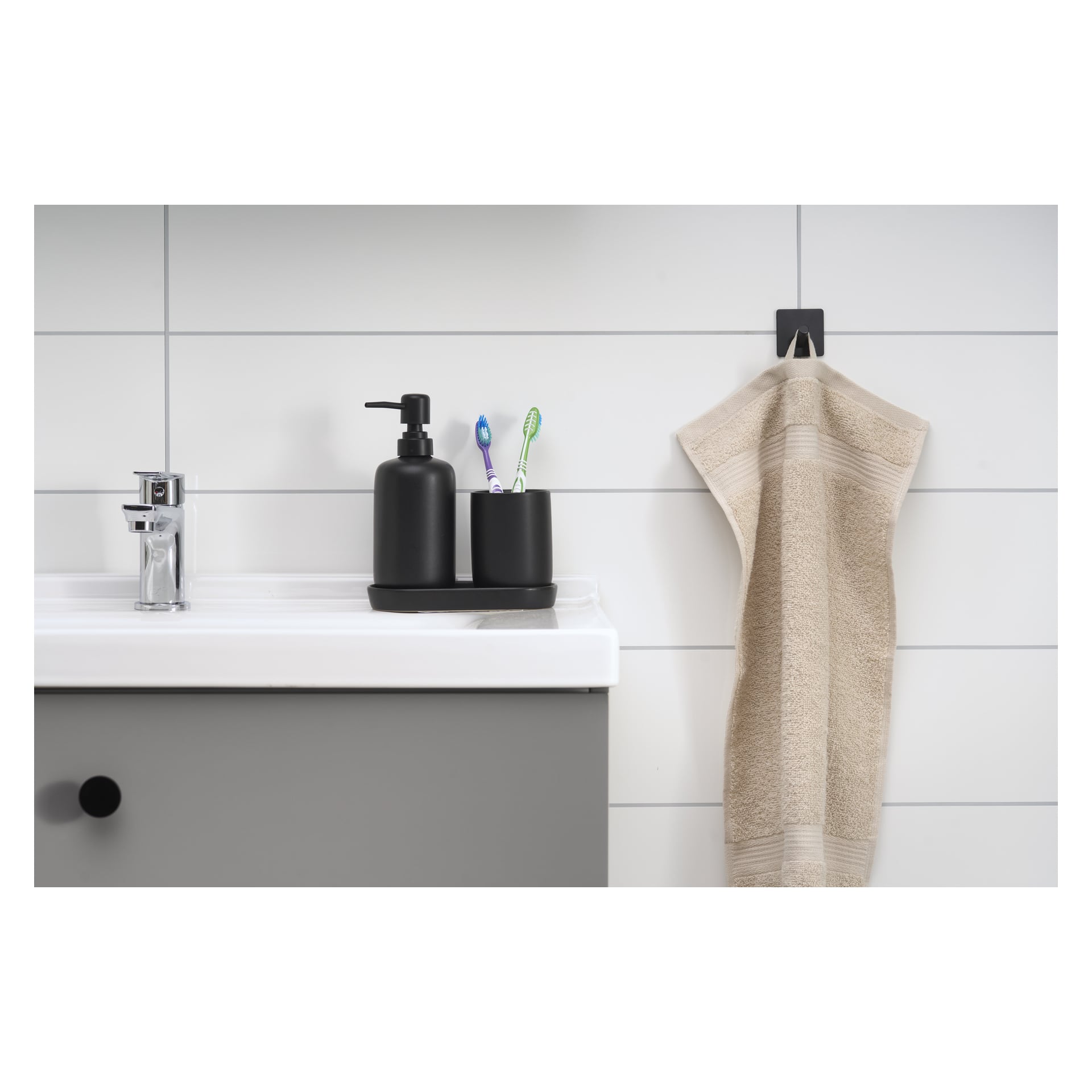 Ett modernt badrumshandfat har en tvålautomat, en hållare med två tandborstar och en kran. Bredvid diskhon hänger en beige handduk från en EKVAP-krok. Scenen utspelar sig mot vita kaklade väggar.