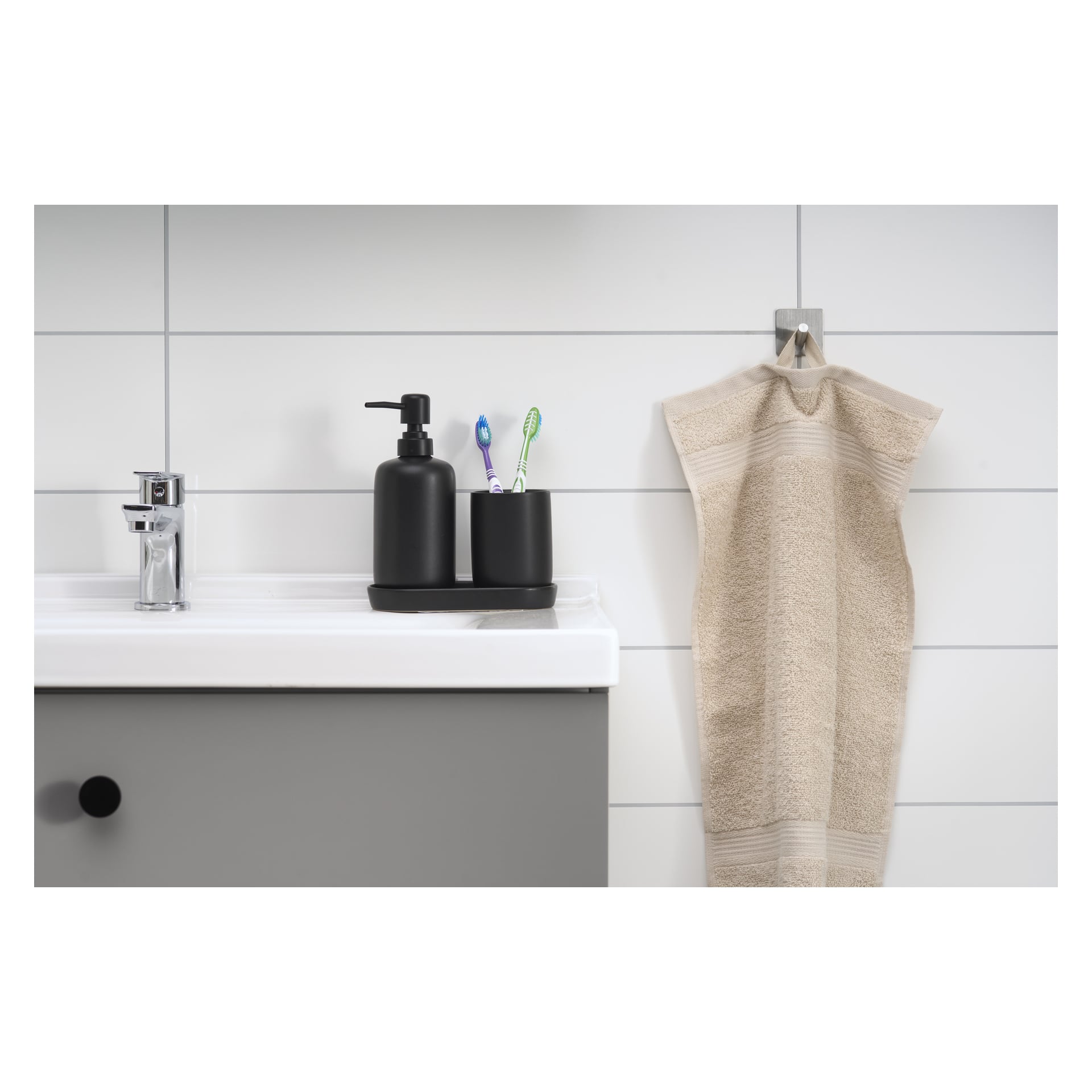 På badrumsbänken finns en handfat med en tvåldispenser och en hållare med tre tandborstar. Bredvid diskhon hänger en beige handduk från en EKVAP-krok på väggen, som är kaklad i vitt.