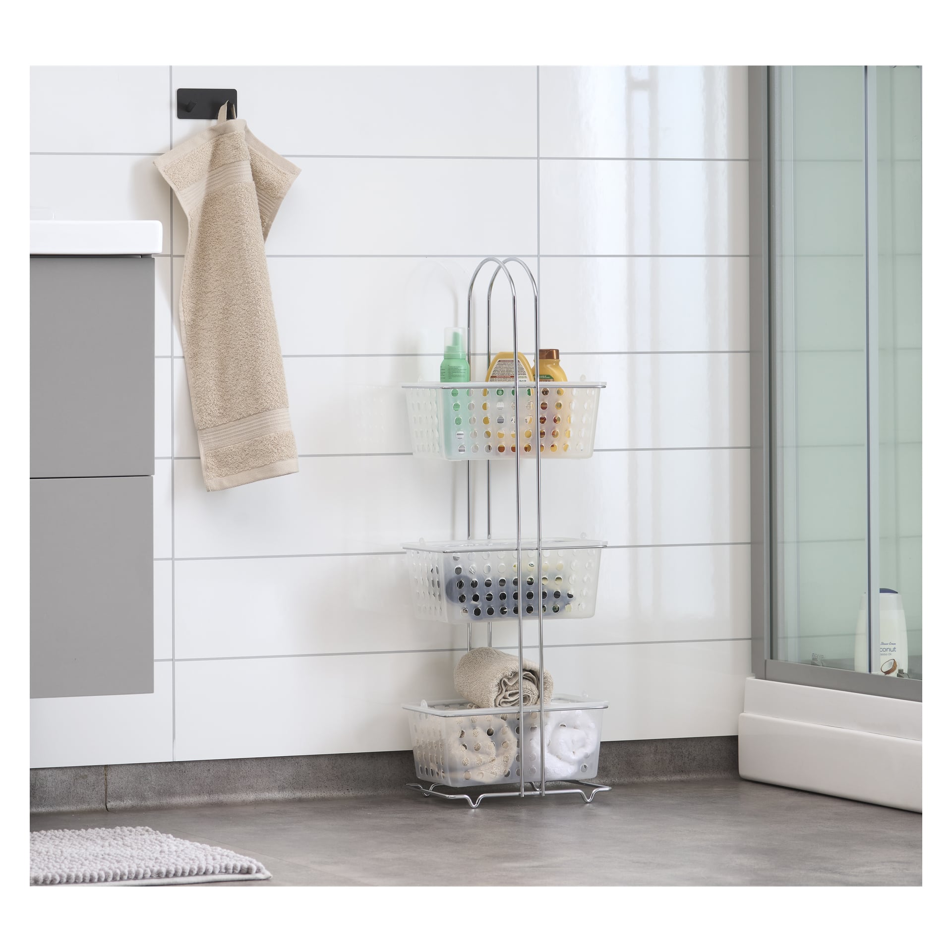 En trevånings EKVIP badrumshylla, med metallhyllor och plastkorgar för att förvara toalettartiklar och handdukar, står på ett kaklat golv bredvid en dusch och en beige handduk hänger i närheten.