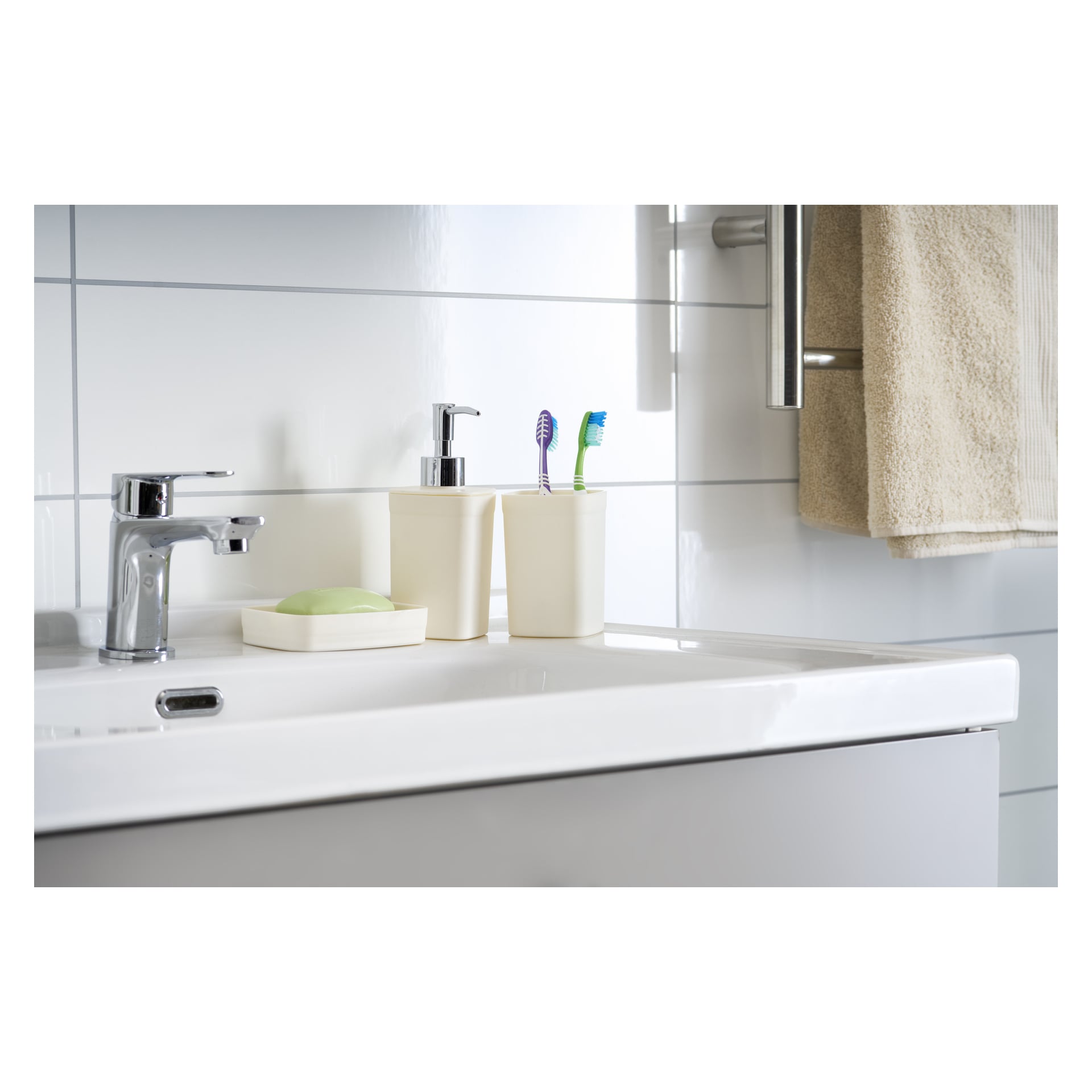 Ett badrumshandfat visar det tredelade EKVIP Soap Dispenser Set, bestående av en kromad tvåldispenser, en matchande tvålskål med grön tvål och en tandborsthållare med två tandborstar. En beige handduk hängs prydligt på en ställning mot vita kakelväggar.