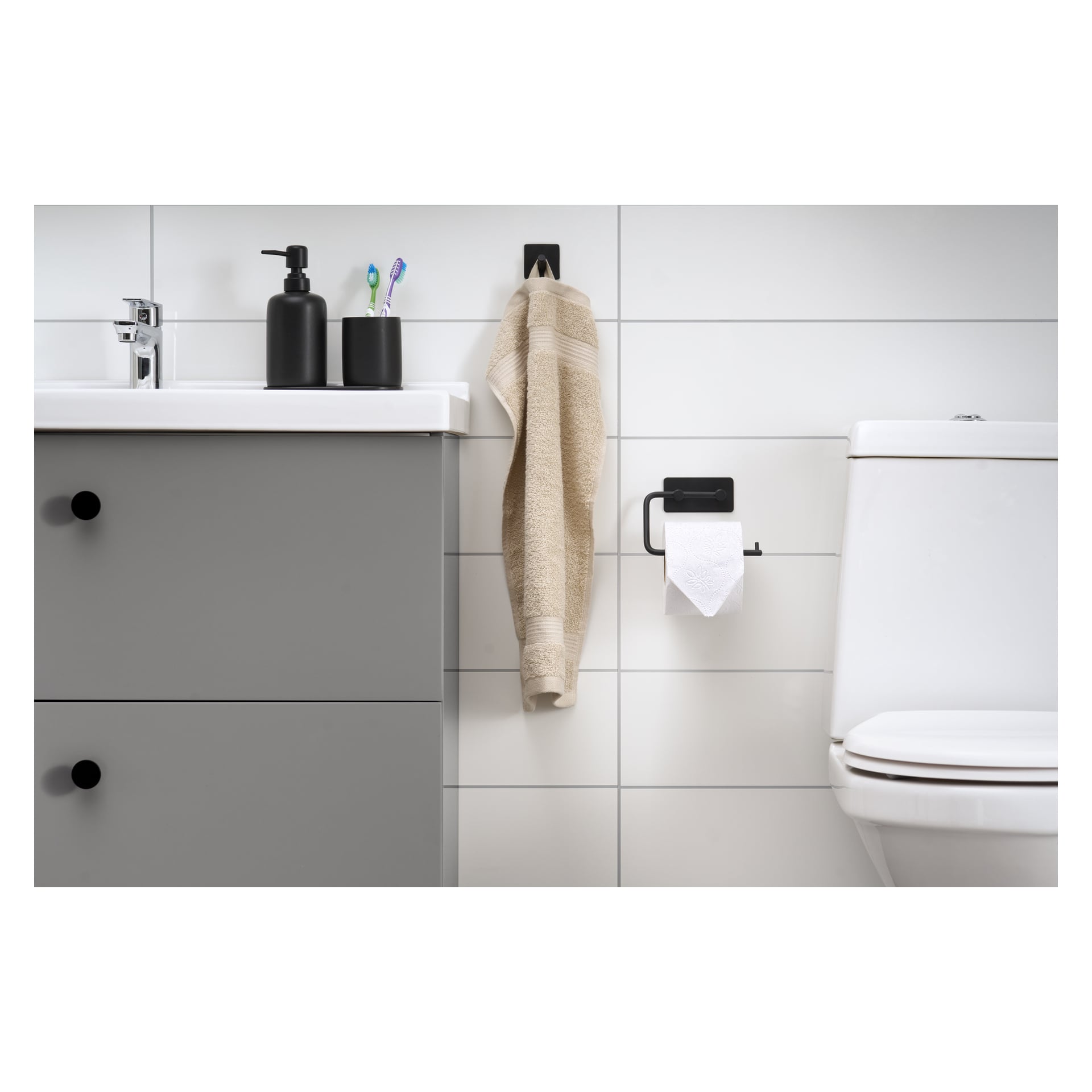 Et stilig bad med en EKVIP toalettpapirholder, grått skap, hvit vask, såpedispenser, to tannbørster i en holder, et beige håndkle hengende på en krok, og et hvitt toalett mot vegger dekket av hvite fliser.