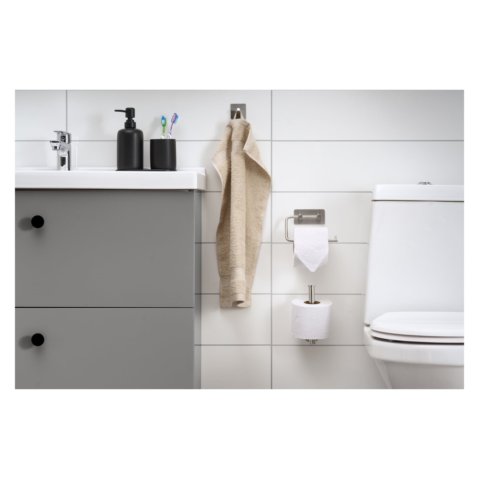 Ett badrum visar EKVIP Spare pappershållaren med två rullar toalettpapper, ett grått skåp, tvålautomat, två tandborstar, en beige handduk och en vit toalett mot vita kaklade väggar.