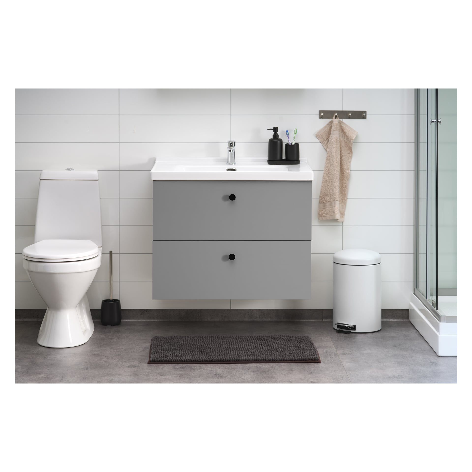 Ett elegant badrum med toalett, grå flytande kommoditet med handfat ovanpå, tvålautomater och tandborstar. En handduk hänger på en krok, medan en vit soptunna och en toalettborste står i närheten. En grå EKVIP badrumsmatta täcker en del av golvet, och det finns en glasinhägnad dusch till höger.