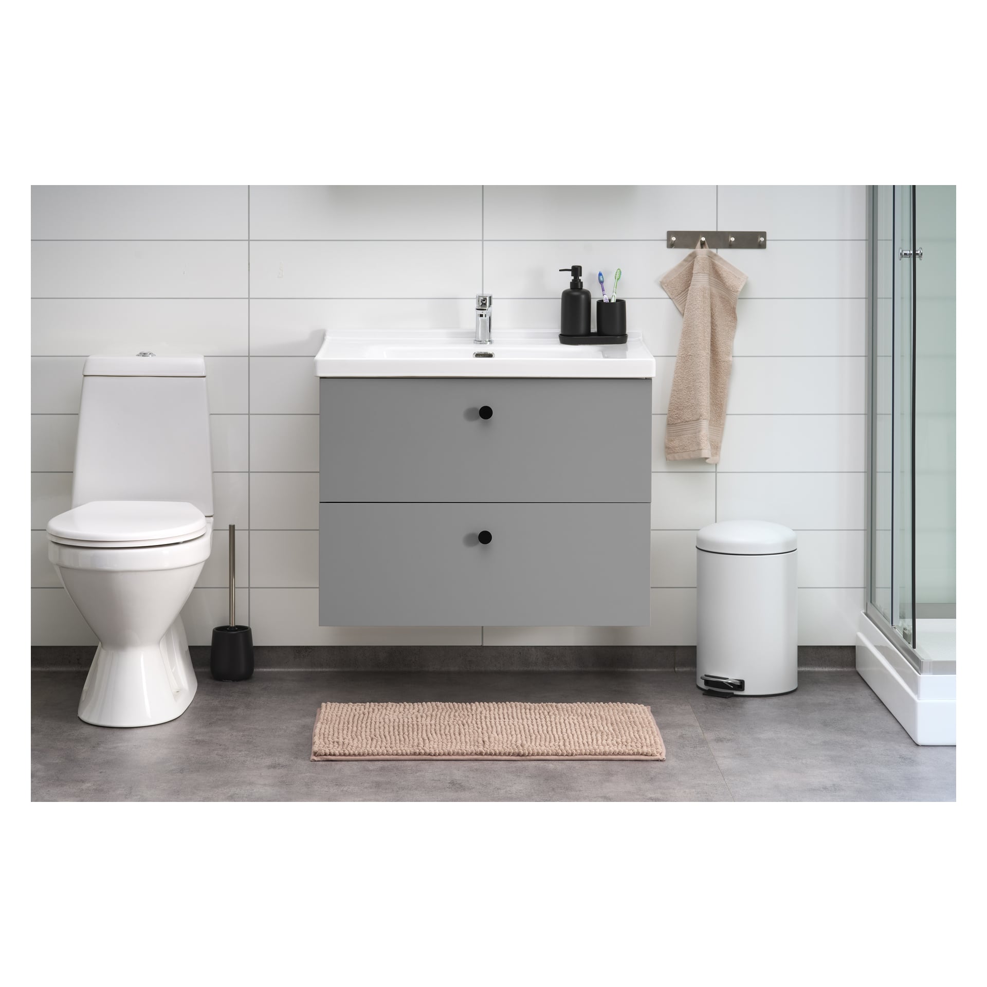Ett stilfullt badrum med en vit toalett, grå flytande sminkbordsduk med handfat ovanpå, hållare för tvål och tandborstar, en beige handduk, vit soptunna, toalettborstset och en rosa EKVIP badrumsmatta på golvet. Ett glasduschutrymme är placerat på höger sida.