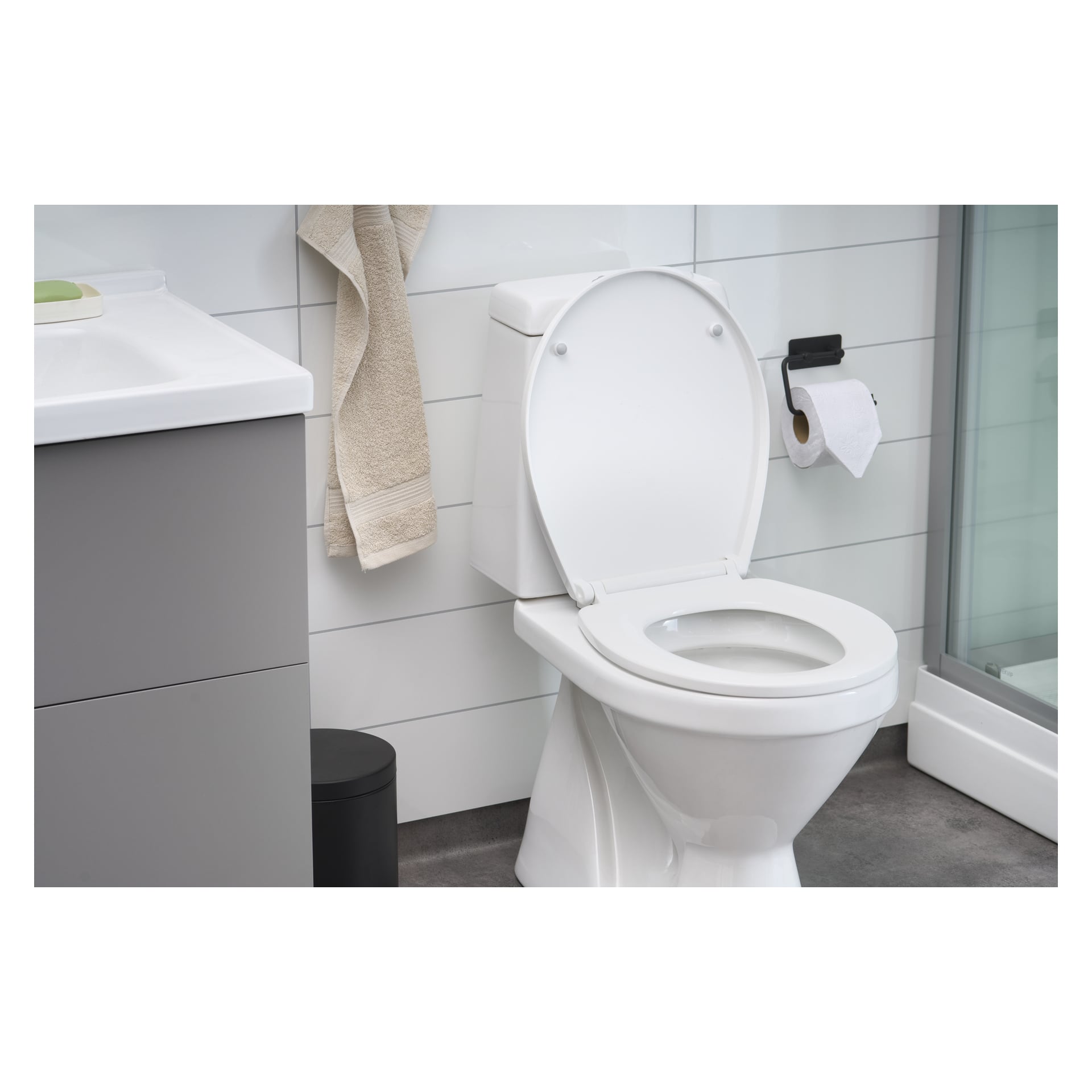 Ett elegant badrum visar upp EKVIP toalettsitsen med det vita locket uppdraget, tillsammans med en handfat, beige handdukar på en krok, toalettpapper på en hållare, en svart soptunna och en glasdörr till duschen i bakgrunden.