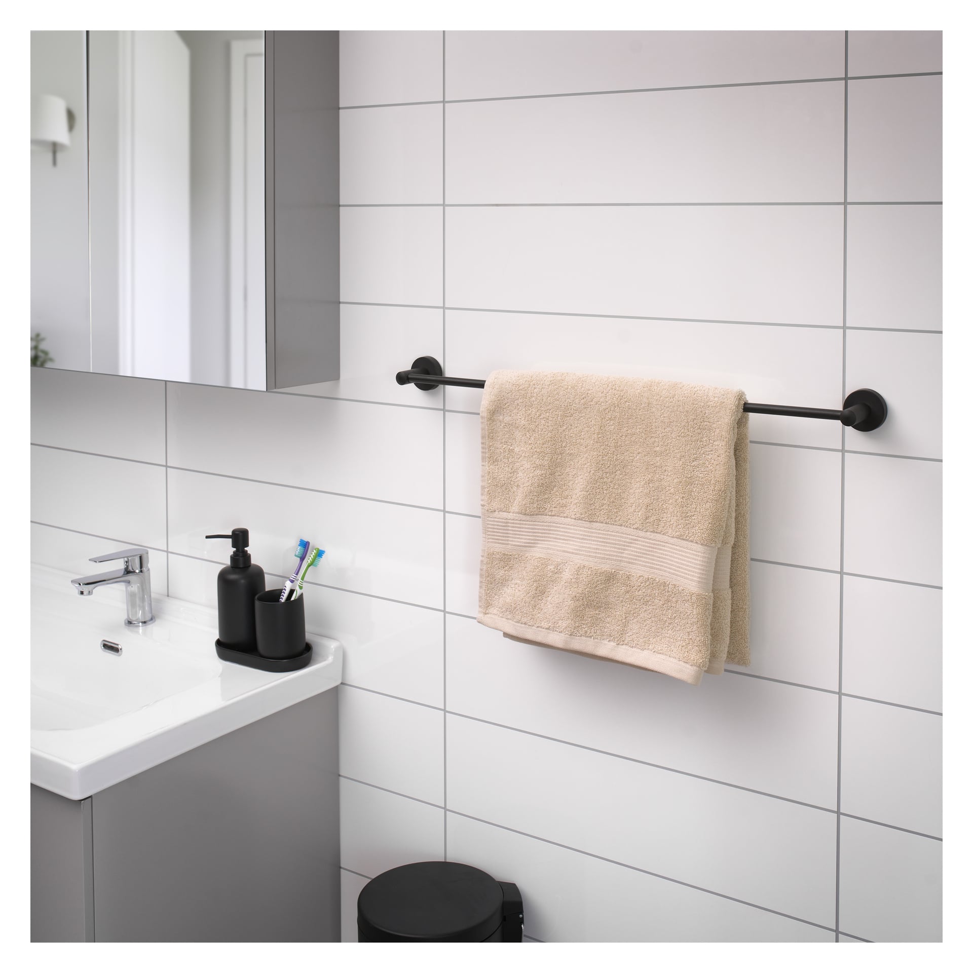 En svart EKVAP-handduksräck är fäst vid en kaklad badrumsvägg och håller en beige handduk. Under den står en svart soptunna. I bakgrunden finns en handfat med en tvålautomat, två tandborstar och en spegel.