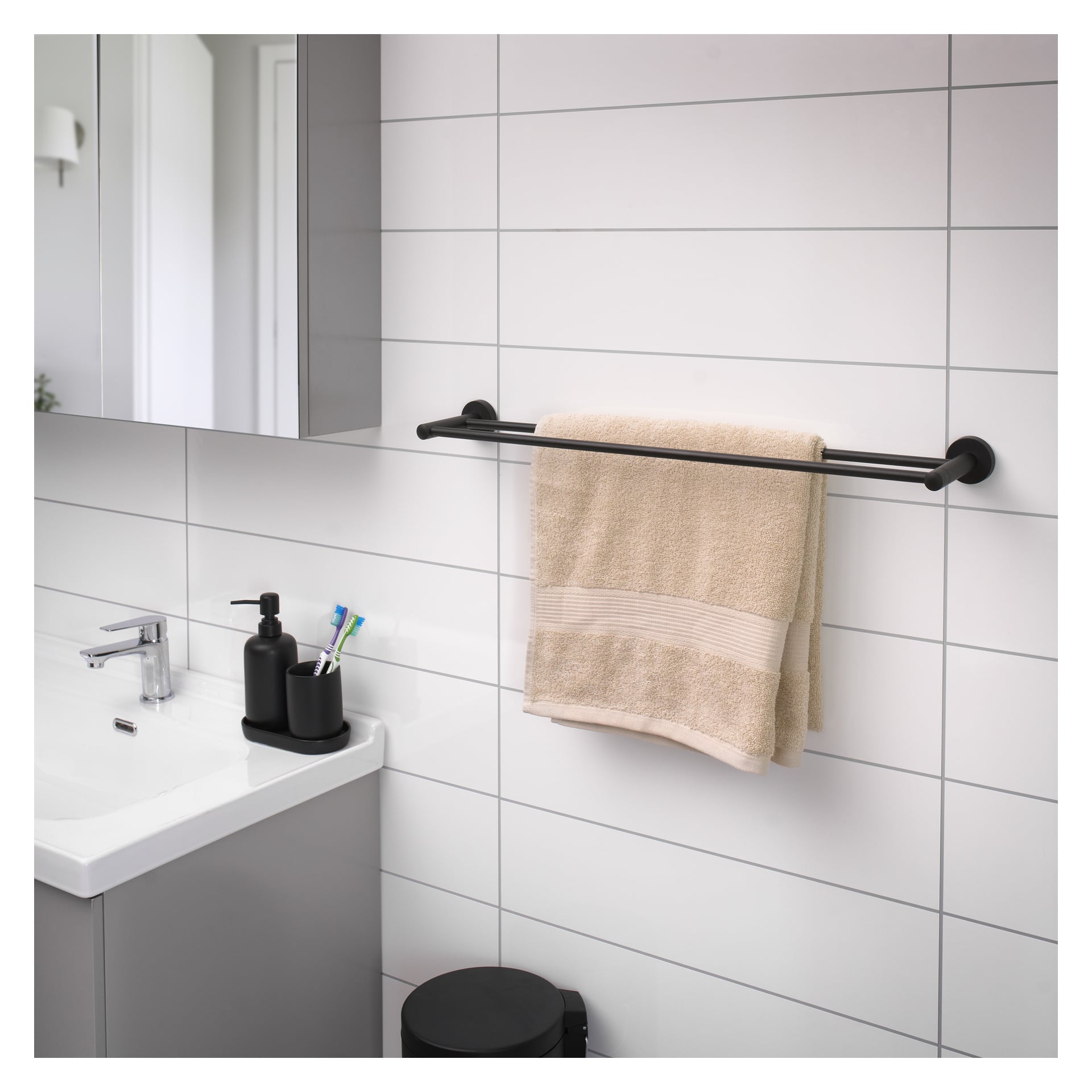 Ett svart EKVAP-handdukshänge är fäst vid en kaklad badrumsvägg och visar en beige handduk. Under finns en vit handfat en svart tvåldispenser och tre tandborstar. En spegel och en del av en dörr syns i bakgrunden.