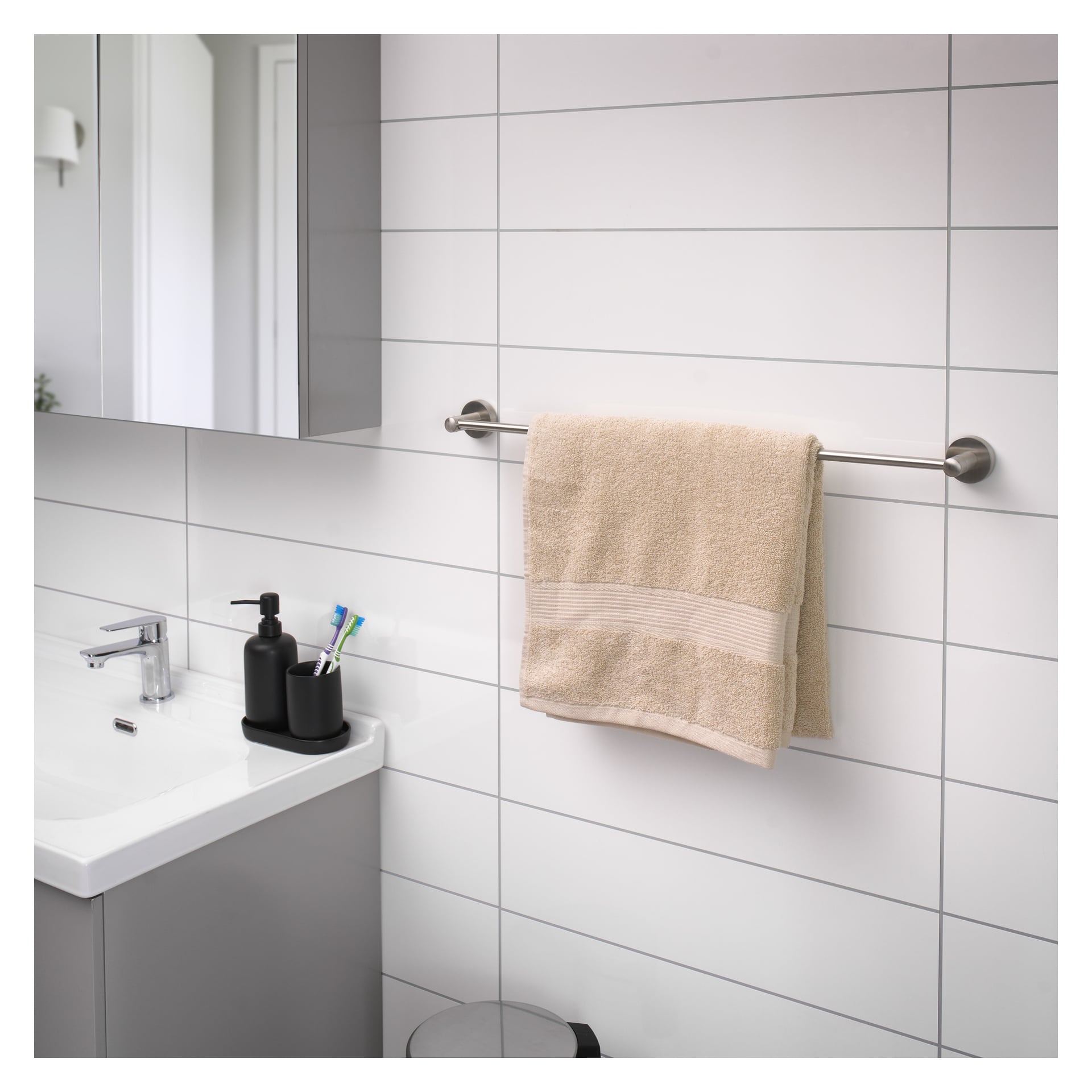 En EKVAP-handduksräck är fäst vid en kaklad badrumsvägg och bär upp en beige handduk. Under räcket finns en handfat med en tvåldispenser och flera tandborstar bredvid en spegel. En del av en soptunna dyker upp längst ner i scenen.