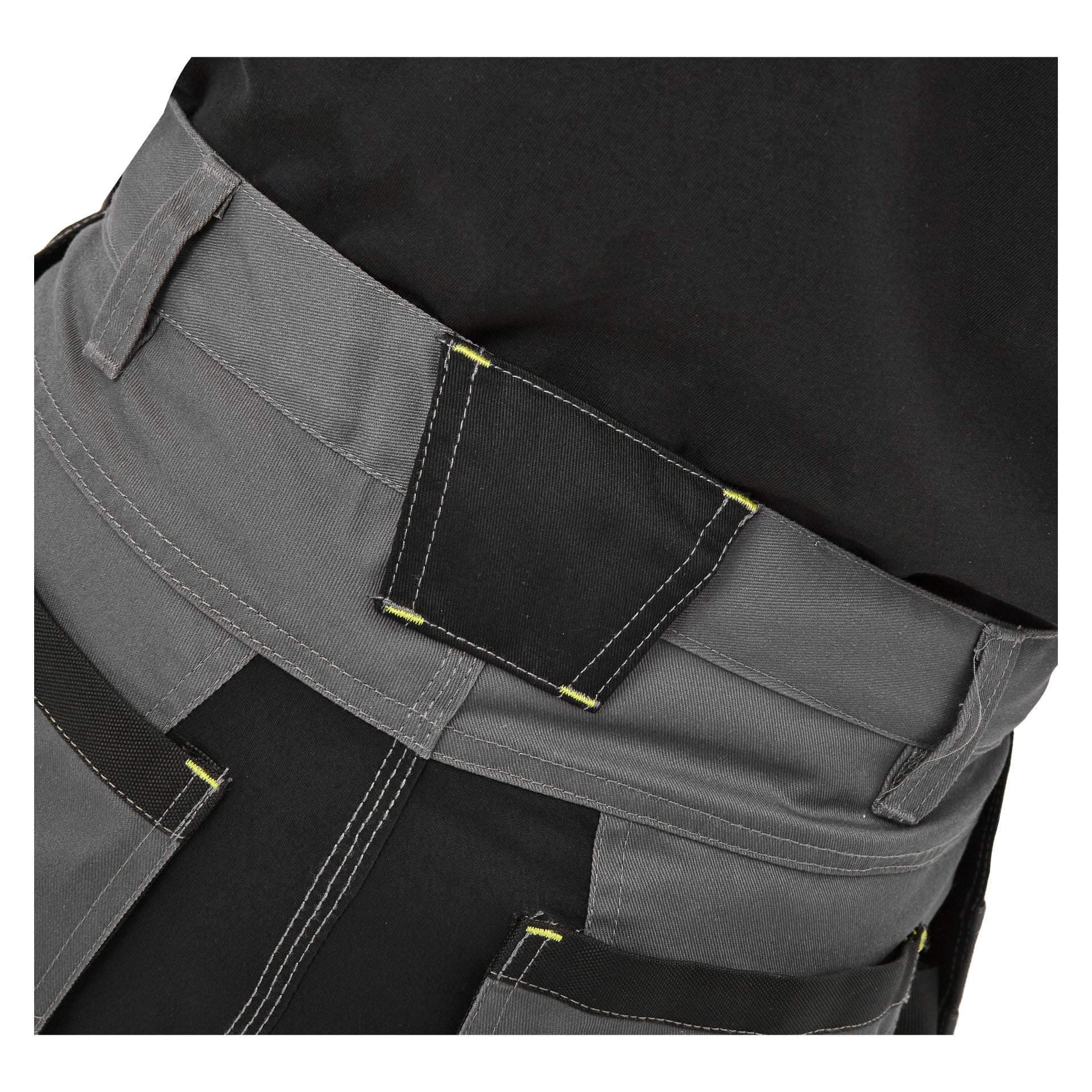 Närbild på en person klädd i Bluewear Men's work shorts Rex, visad i grått och svart med förstärkt söm och flera synliga fickor, tillsammans med en svart skjorta instoppad i midjebandet. Fokus ligger på det robusta tyget och den robusta konstruktionen.