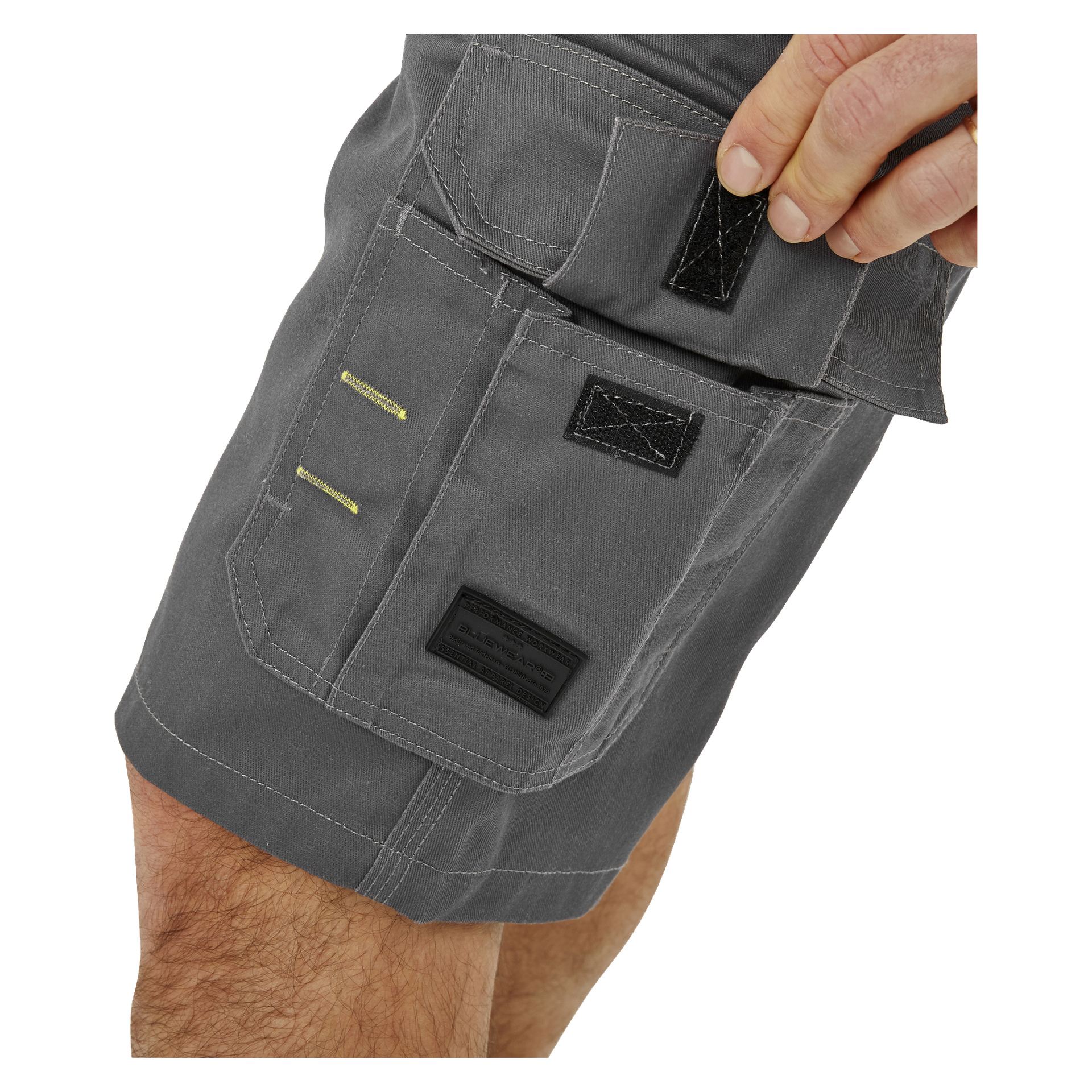En persons hand lyfter kardborrebandsfliken på fickan på grå Bluewear Men's Work Shorts Rex, vilket visar de intrikata sömmen och en svart rektangulär etikett på fickan. Endast handen och en del av ett ben visas.