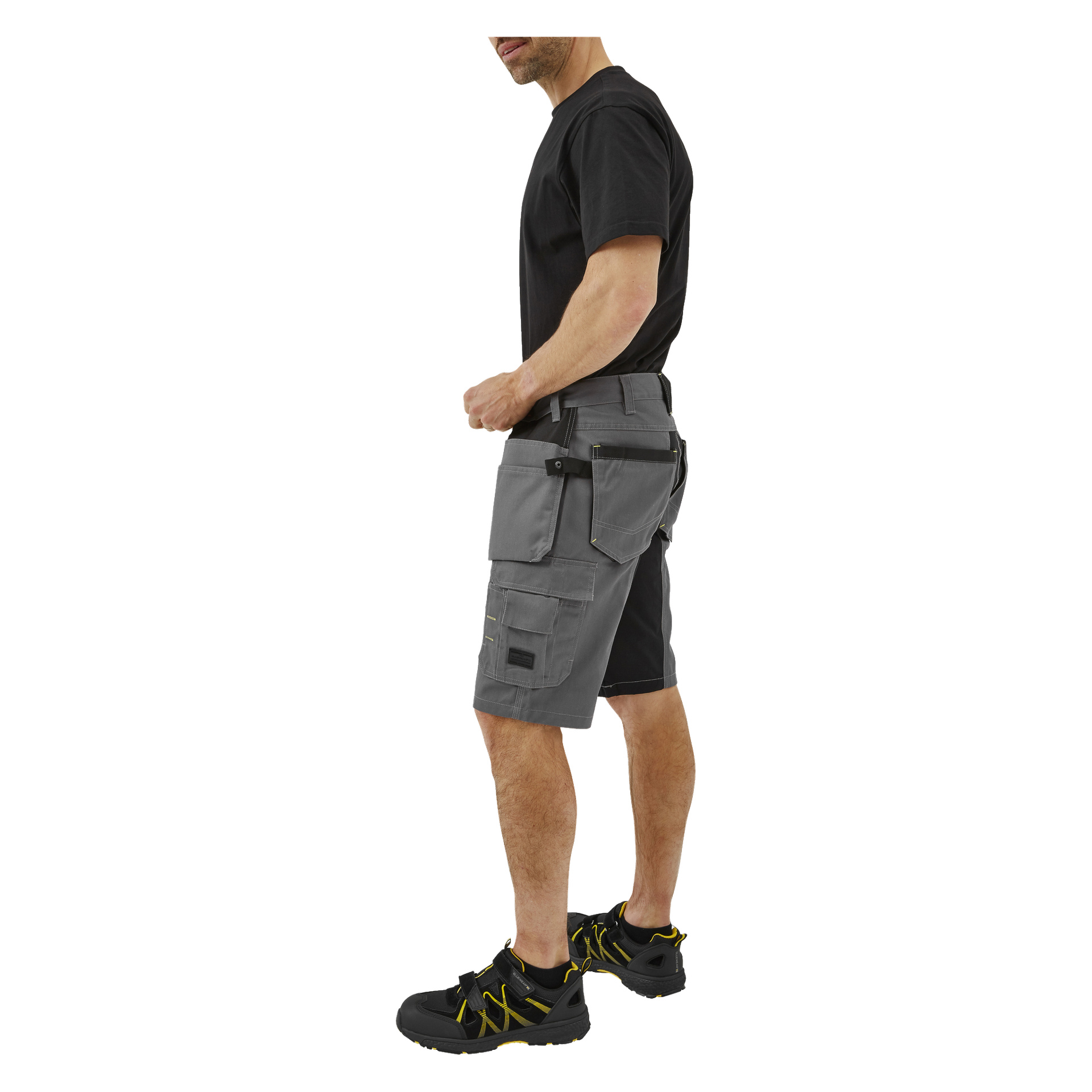 En man visas från huvud till mitten av vaden, stående på sidan. Han bär en svart t-shirt, grå Bluewear Men's Rex arbetsshorts med verktygsfickor, svart bälte och svarta och gula arbetsskor.