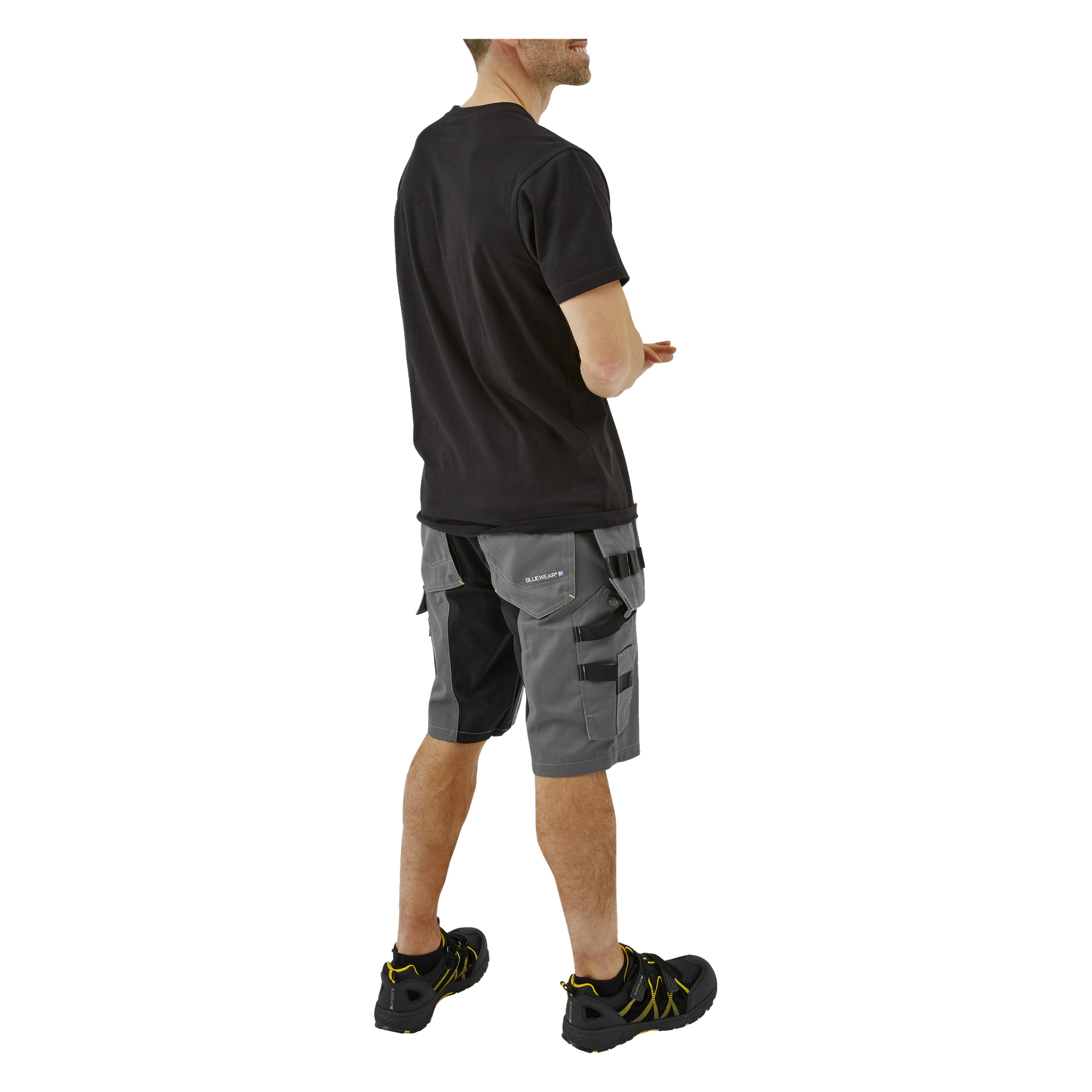 En man syns bakifrån, klädd i en svart t-shirt, grå Bluewear Workshorts för herrar med fickor och svarta arbetsskor med gula detaljer. Hans armbågar är lätt böjda.