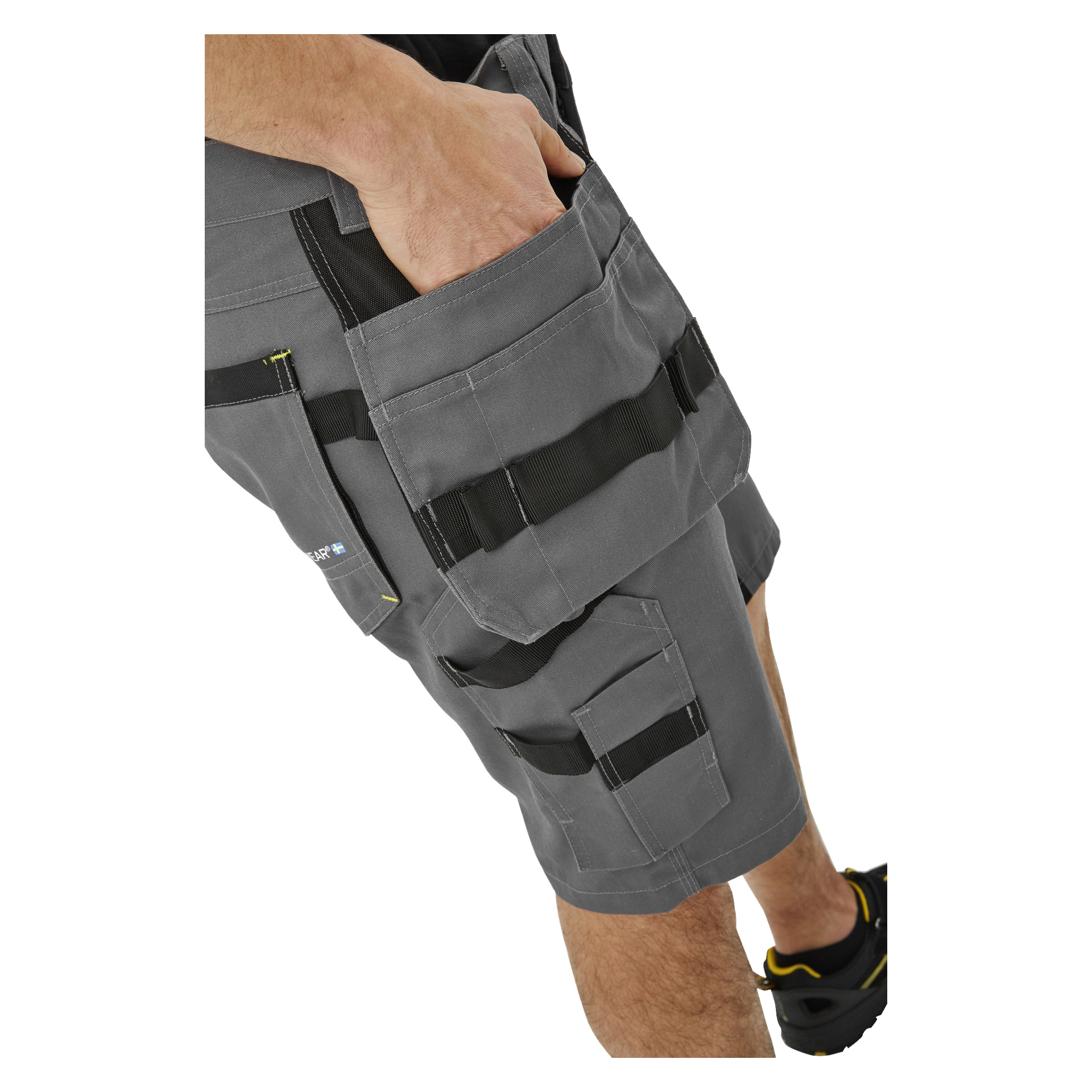 Bilden visar en person från midja till knän som bär Bluewear Men's Rex arbetsshorts, som har svarta remmar och flera fickor, med en hand vilande inuti en ficka.