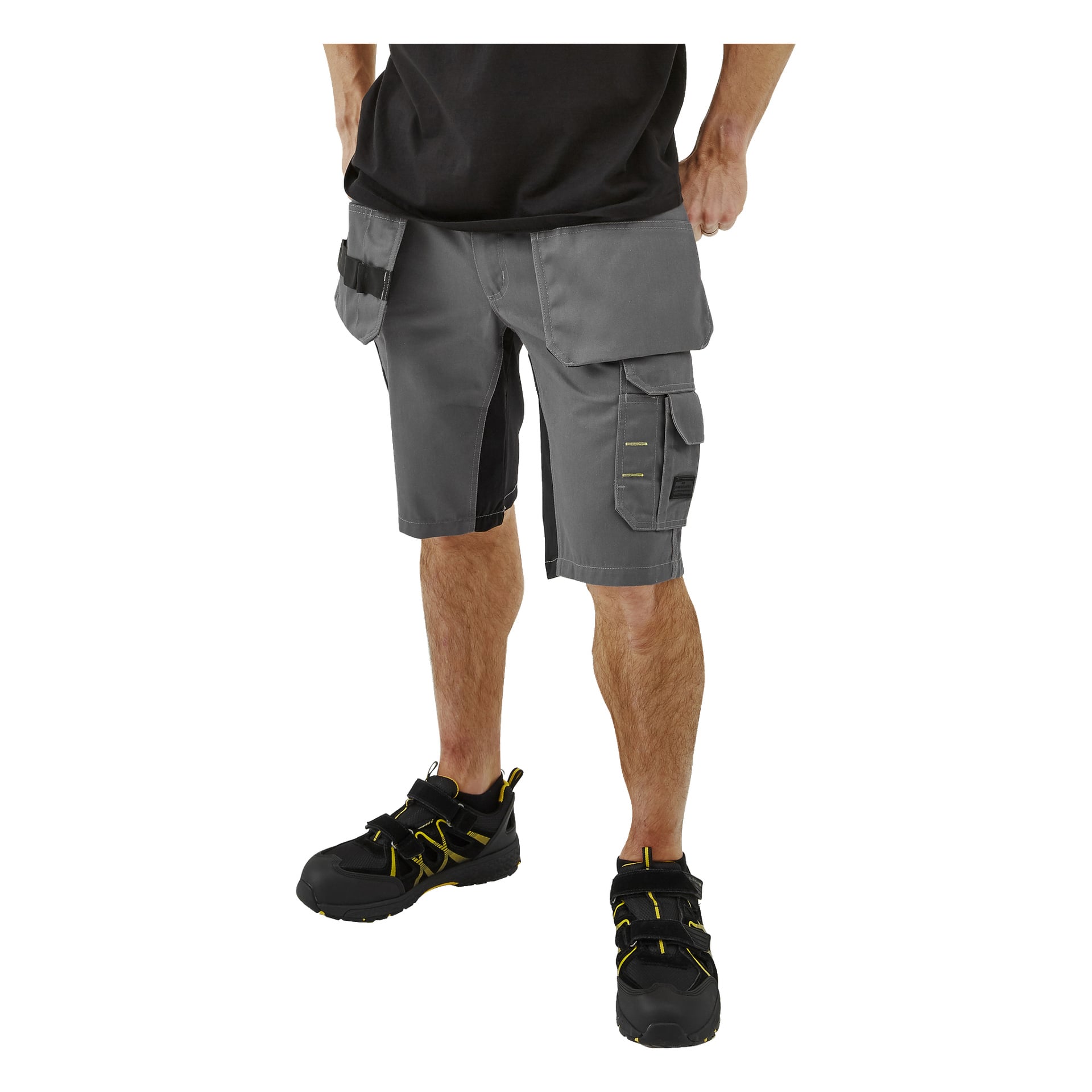 En person visas från midjan och neråt, klädd i Bluewear Men's Rex arbetsshorts med cargofickor och svarta arbetsskor framhävda av gula detaljer, stående med händerna på höfterna.