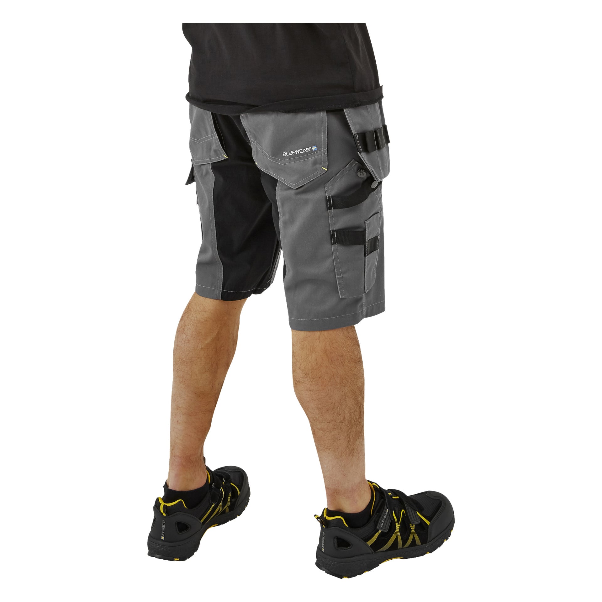 En person i svart skjorta, grå Bluewear Men's Rex arbetsshorts med svart kant och fickor samt svarta sandaler med gula detaljer står vänd bort från betraktaren.