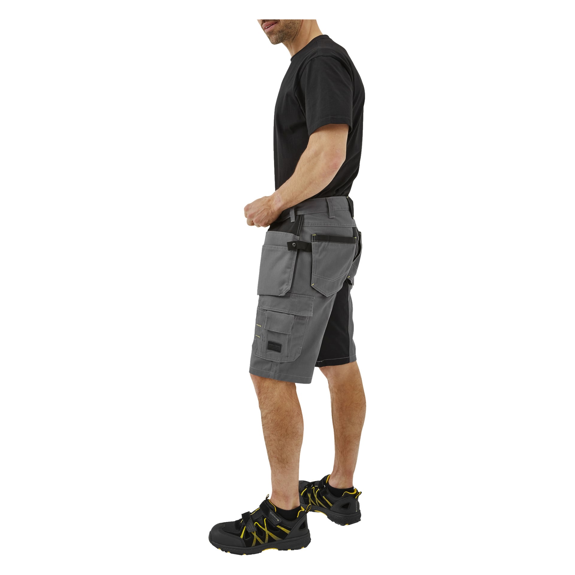 En man står i profil iklädd en svart t-shirt, grå Bluewear Men's work shorts Rex med flera fickor, svart bälte och svarta och gula arbetsskor. Hans vänstra hand är placerad nära höften.