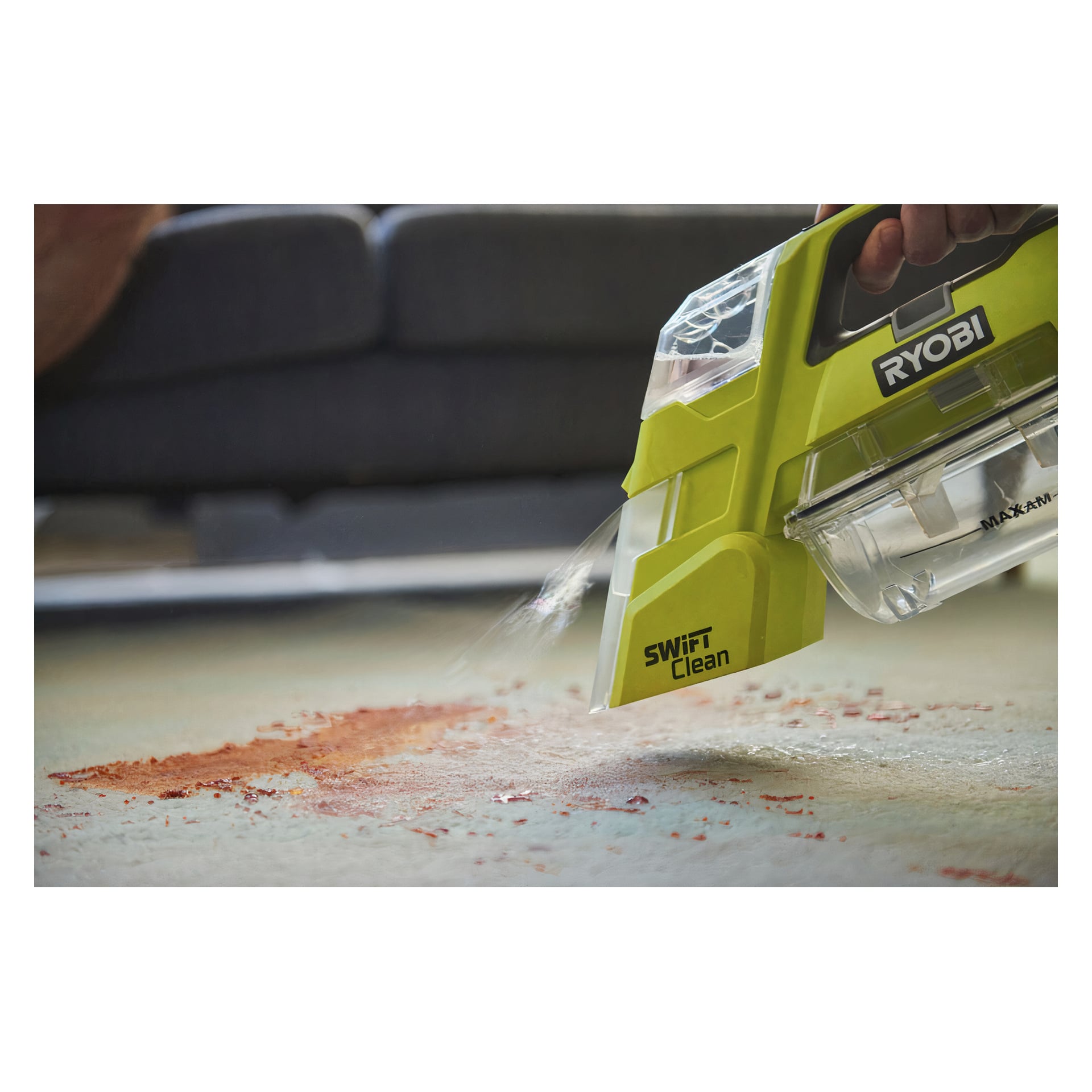 Käsi ohjaa Ryobi One+ Swift Clean RBCLS-01 -paristokäyttöistä tahranpoistoainetta puhdistaakseen matolta vuotaneen jauhemaisen aineen, taustalla pehmeästi sumentuneena sohva.