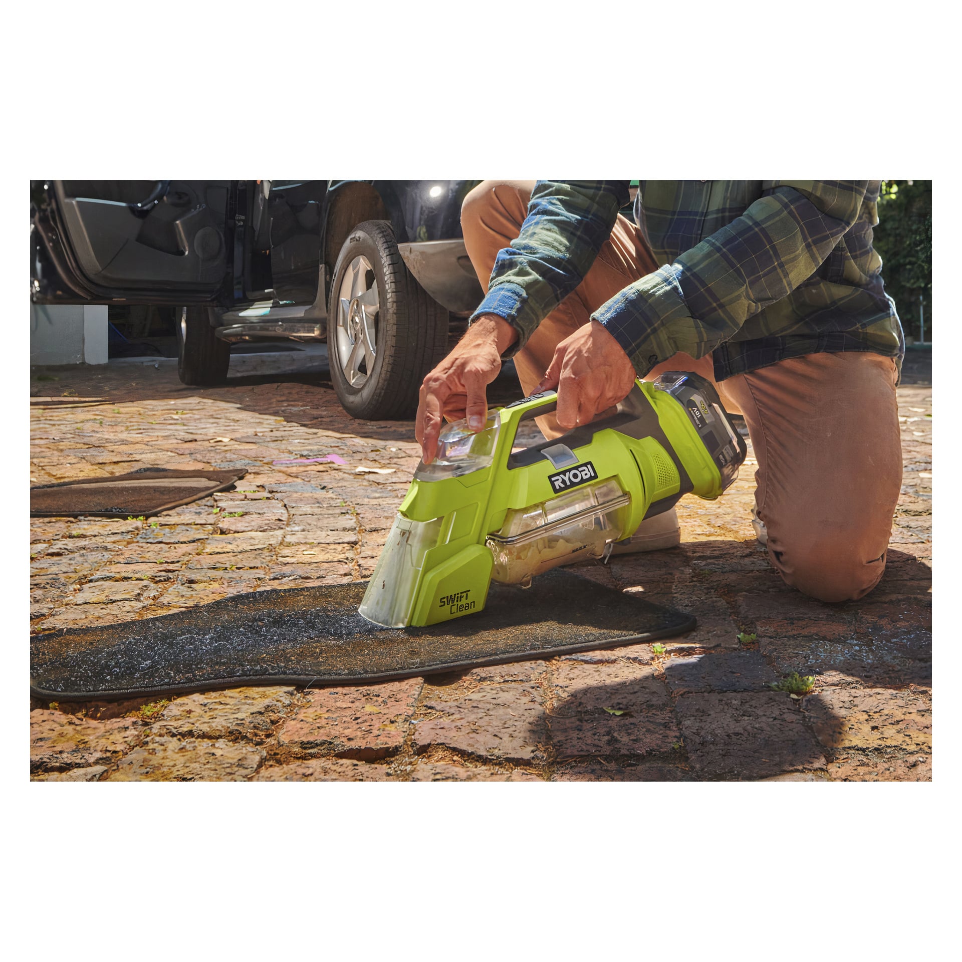 Polvistuneena tiilipihalla henkilö käyttää Ryobi One+ -paristokäyttöistä tahranpoistoainetta Swift Clean RBCLS-01 mustaa auton lattiamattoa. Taustalla on avoin auton ovi ja toinen matto.