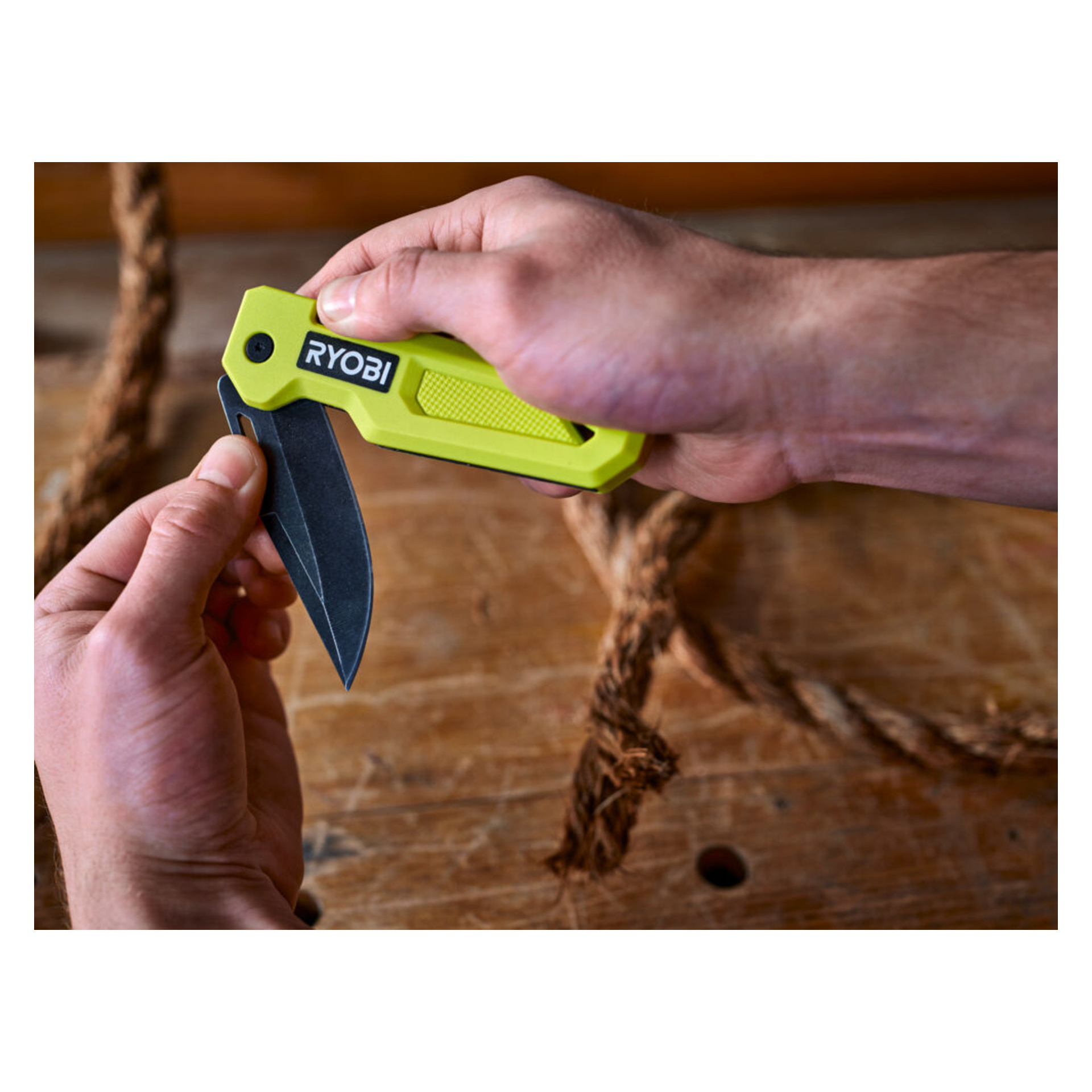 En person öppnar en grön Ryobi Jack knife RHCKF-3 på en träyta, med ett hoprullat rep synligt i bakgrunden.